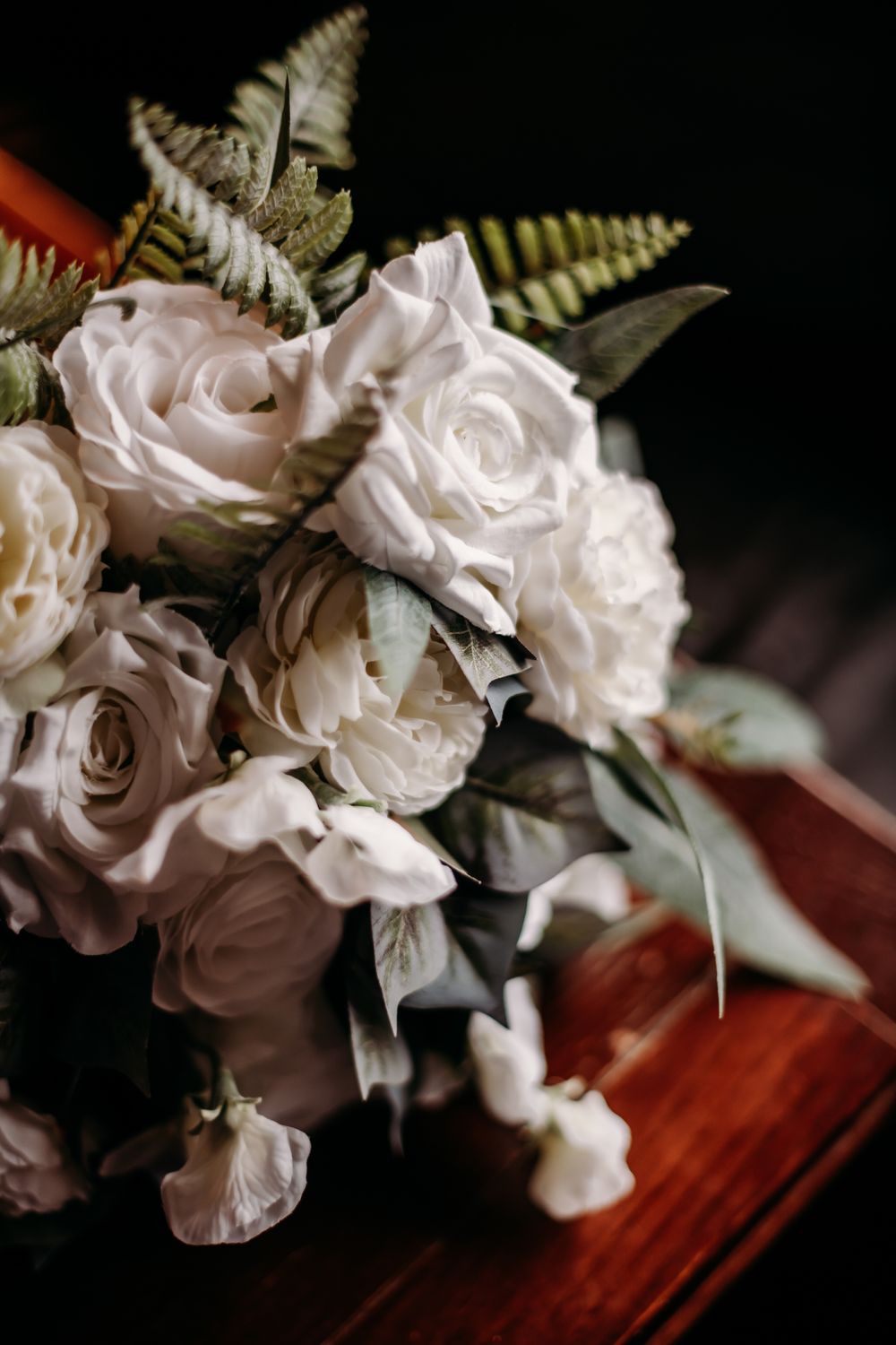 A moody and elegant wedding bouquet of white roses with fern leaves rests on a dark wooden surface.