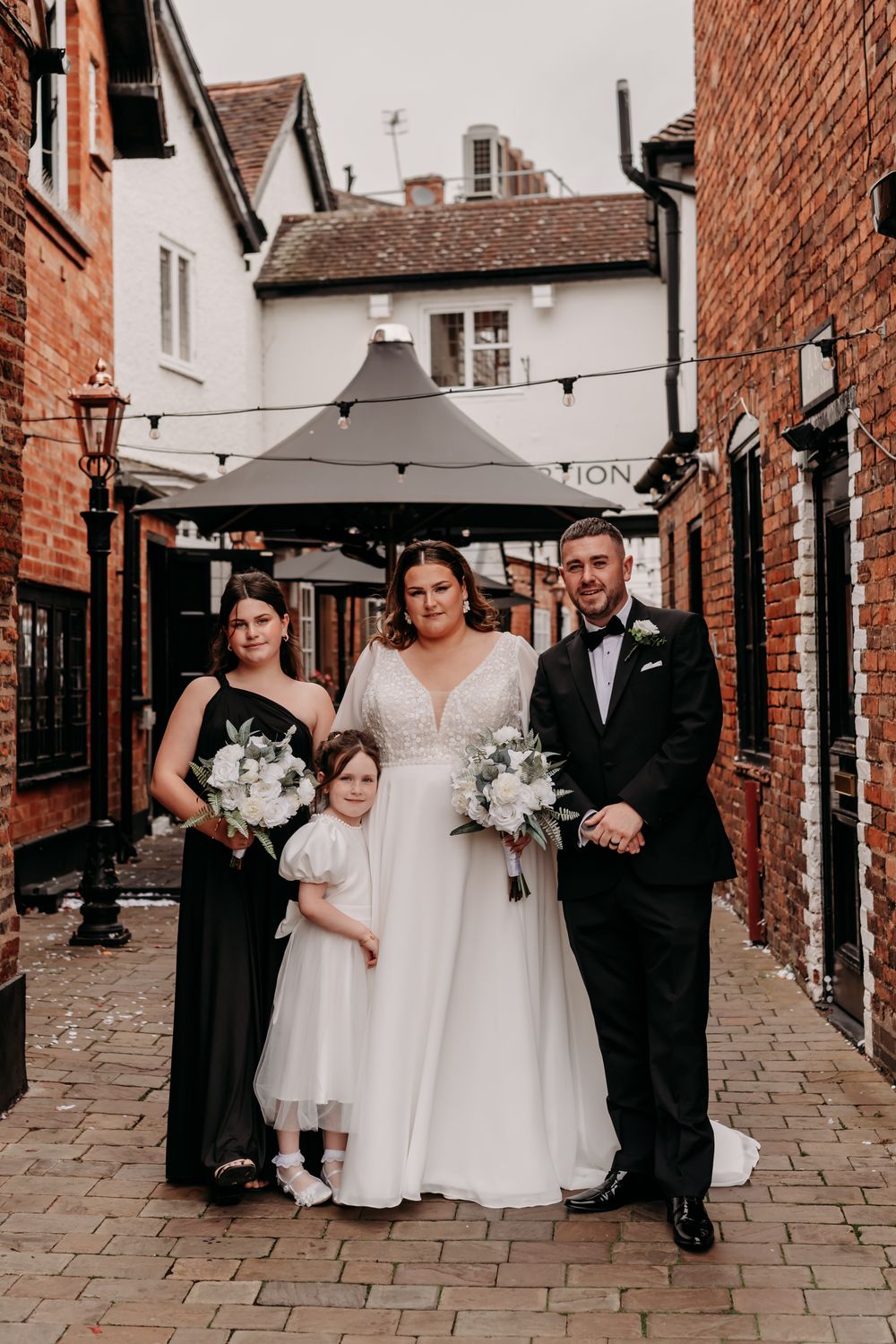 Wedding party walking down cobblestone alley between brick buildings with black umbrellas on a rainy day.