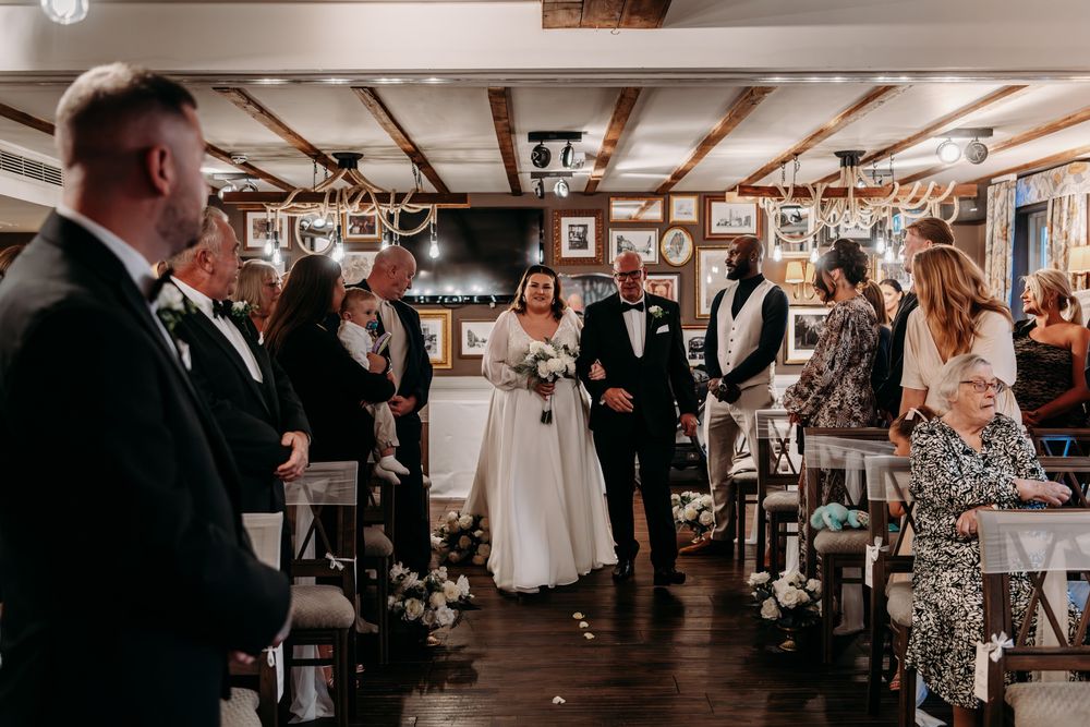 Wedding reception guests gather on a rustic dance floor with wooden beams and ambient lighting overhead.