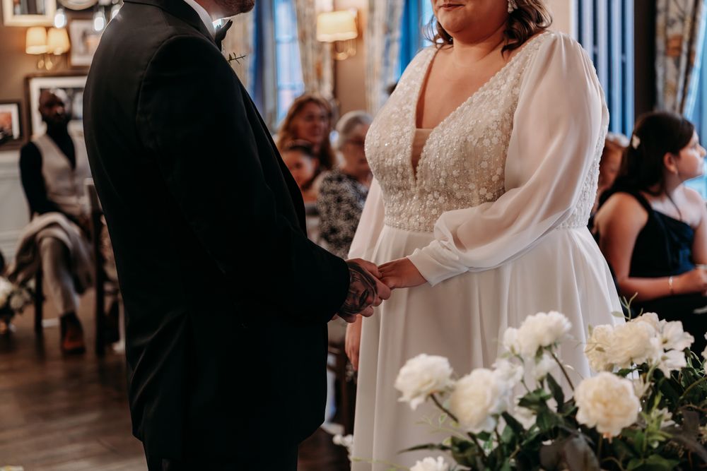 A couple holds hands during their wedding ceremony with white floral arrangements in the foreground.