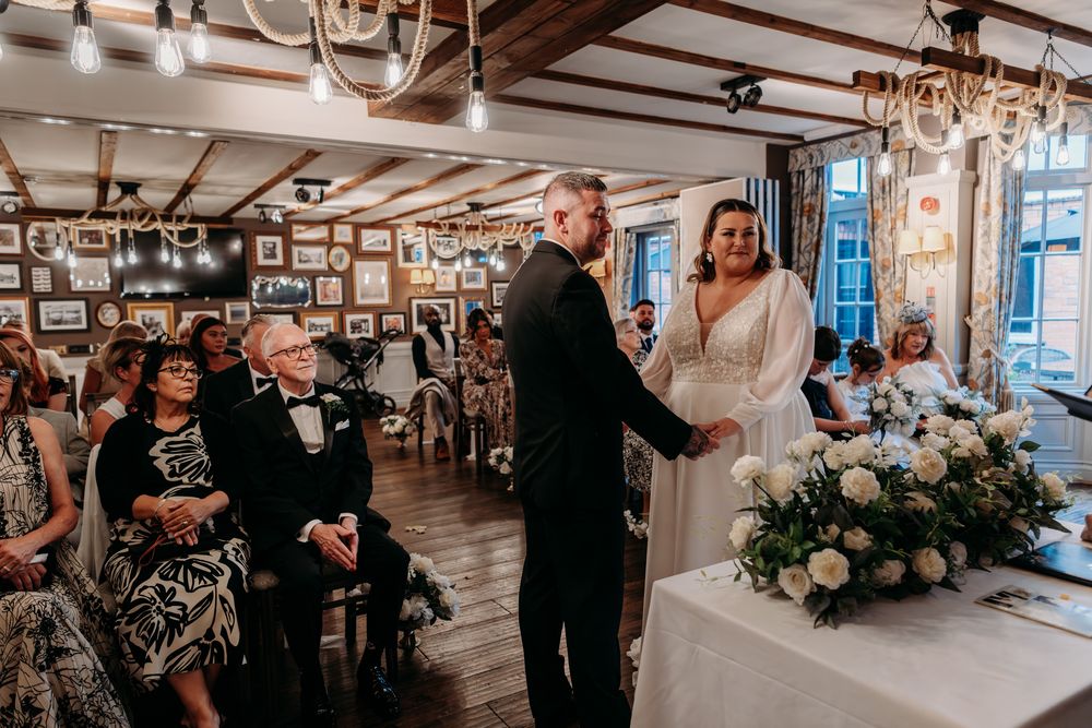 A wedding ceremony takes place in a rustic indoor venue with wooden beams and elegant chandeliers.