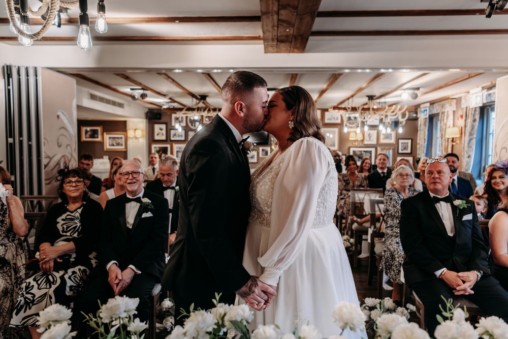 A couple shares a romantic moment during their wedding ceremony surrounded by guests and floral arrangements.