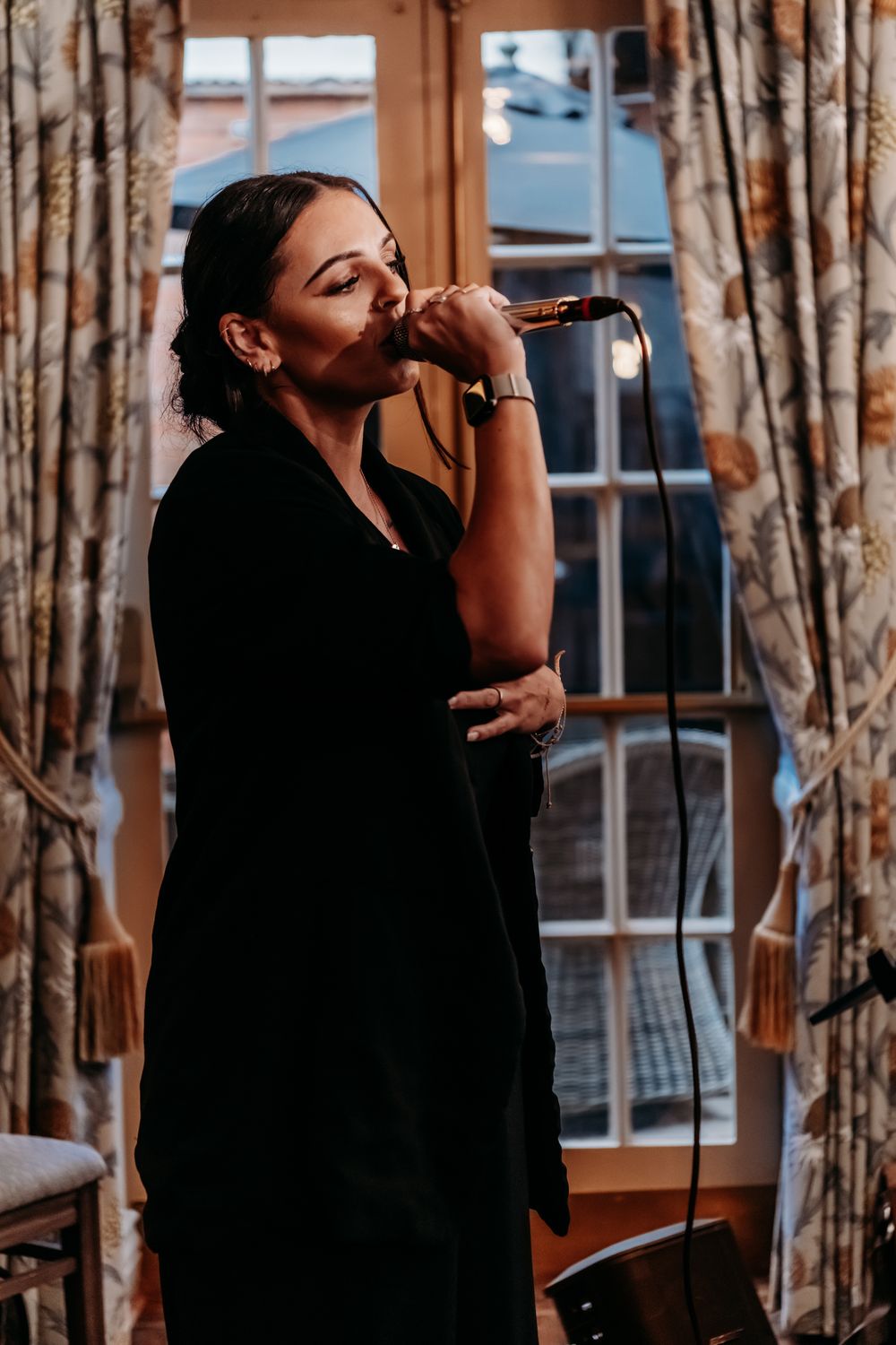 A performer in black clothing sings emotionally into a microphone during a live performance against patterned curtains.