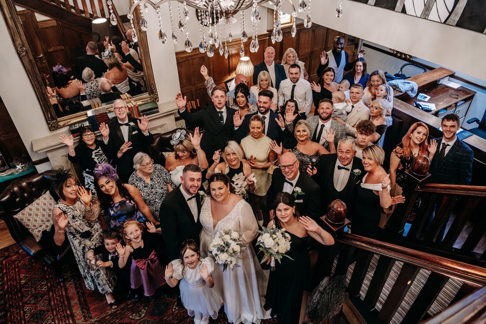 Wedding guests gather in a reception hall to celebrate with joyful expressions and raised hands.