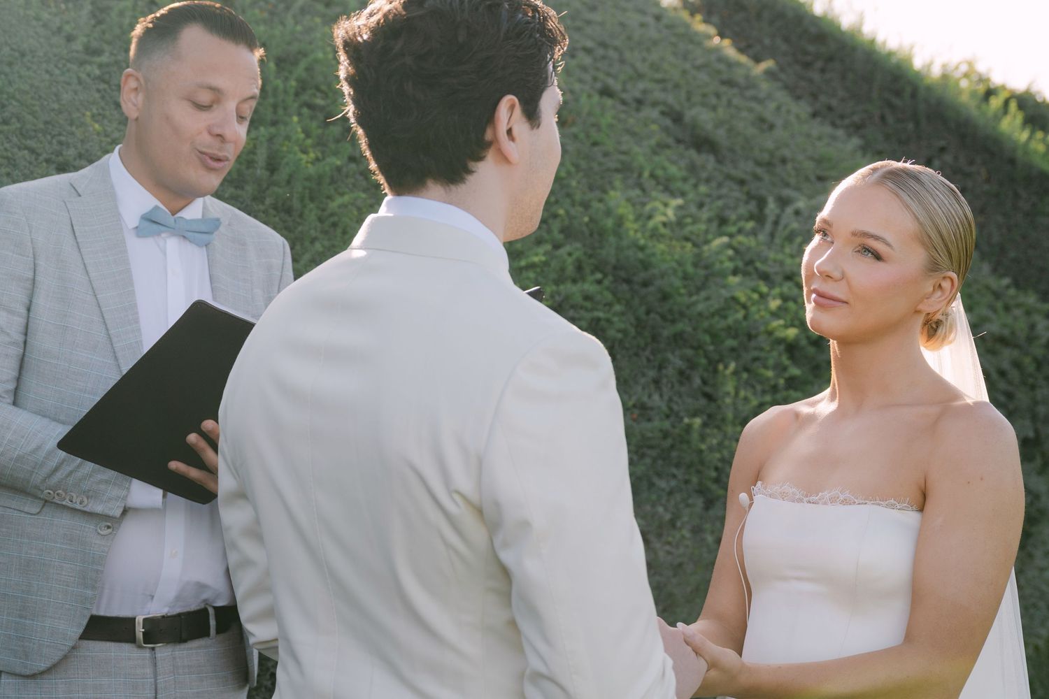 A couple stands together during an outdoor wedding ceremony surrounded by greenery and guests.