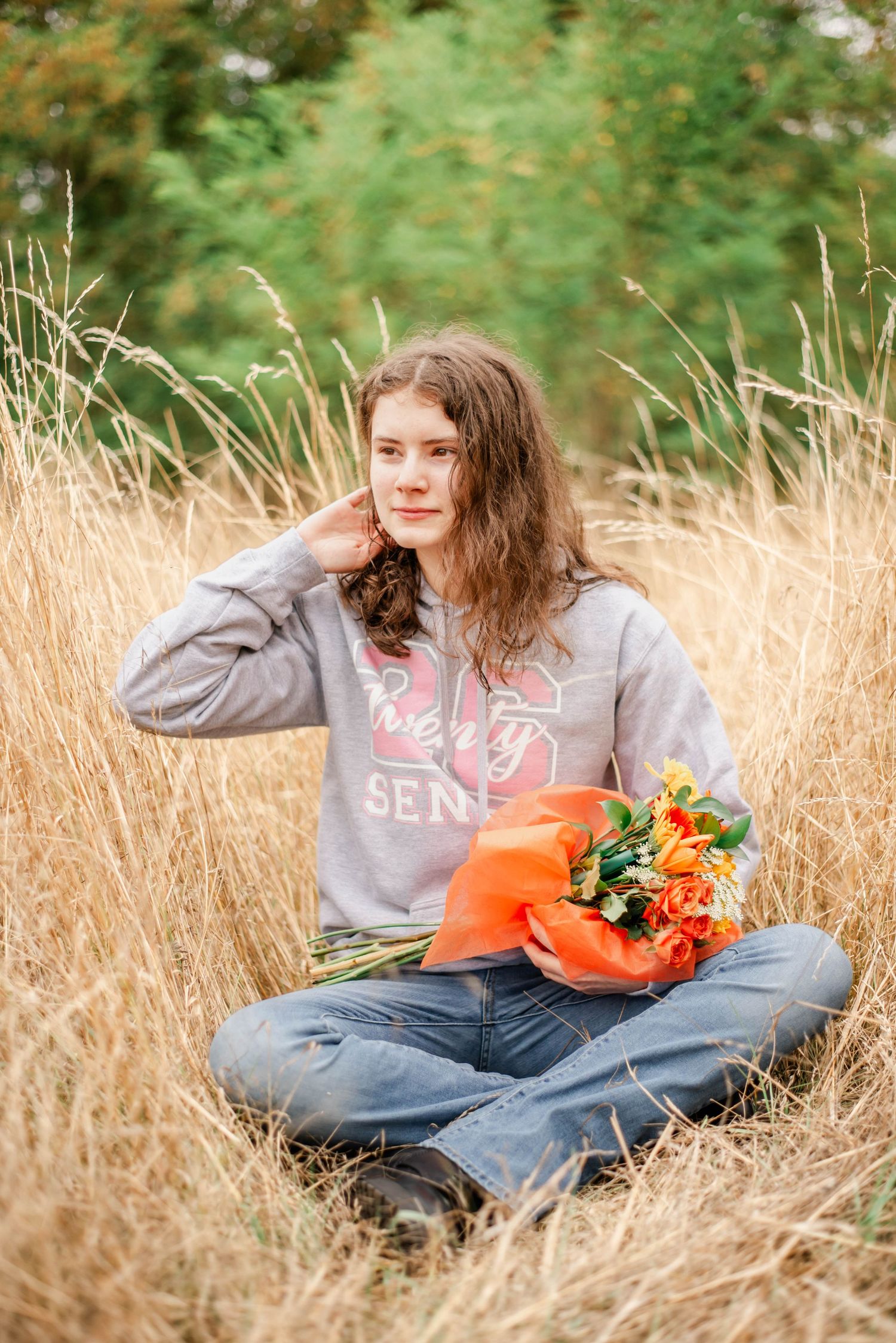 A person in a gray sweatshirt sits in tall golden grass holding an orange pumpkin with flowers.