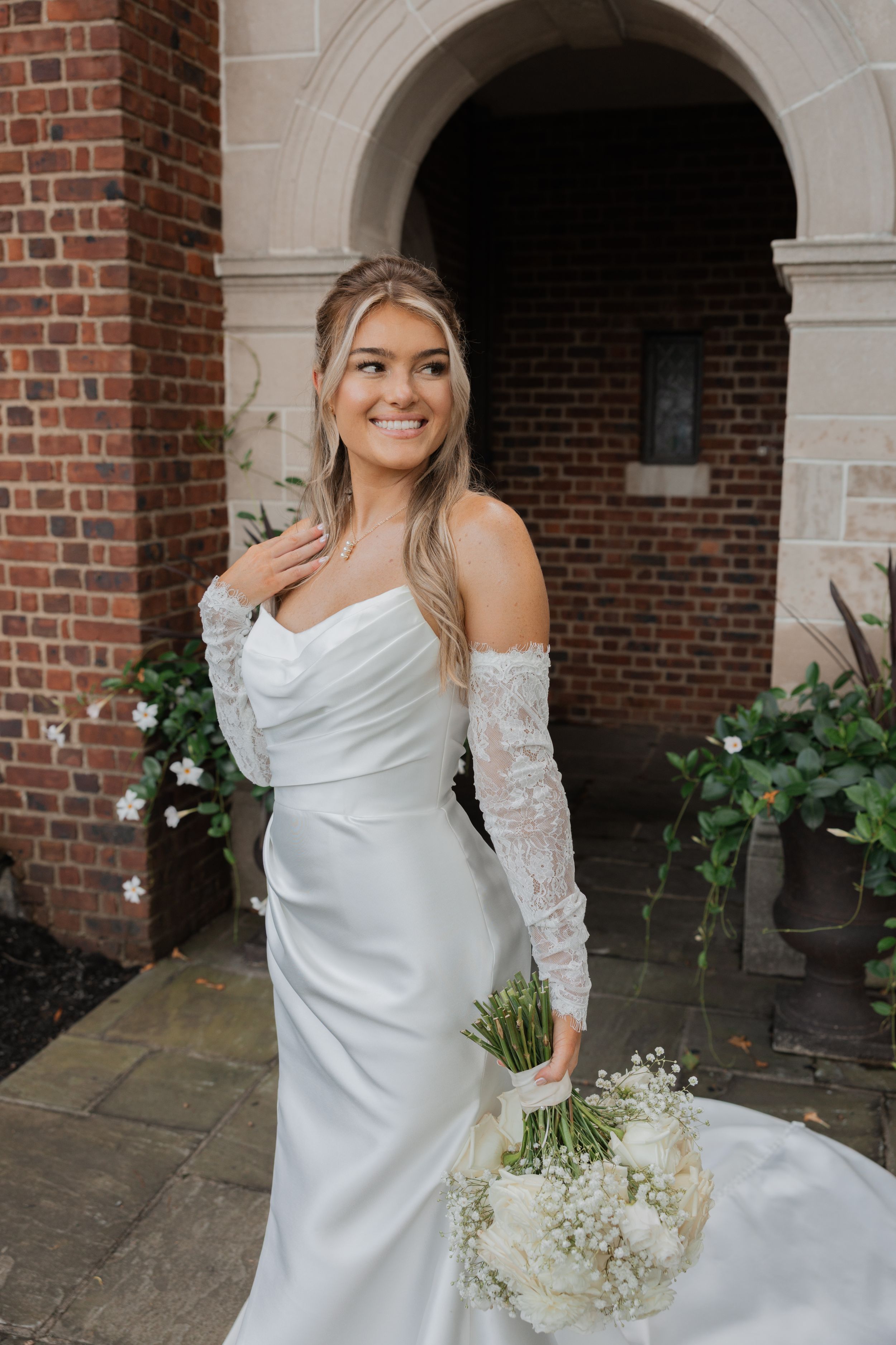 Bride in elegant white off-shoulder gown with lace sleeves poses against brick archway holding white floral bouquet.