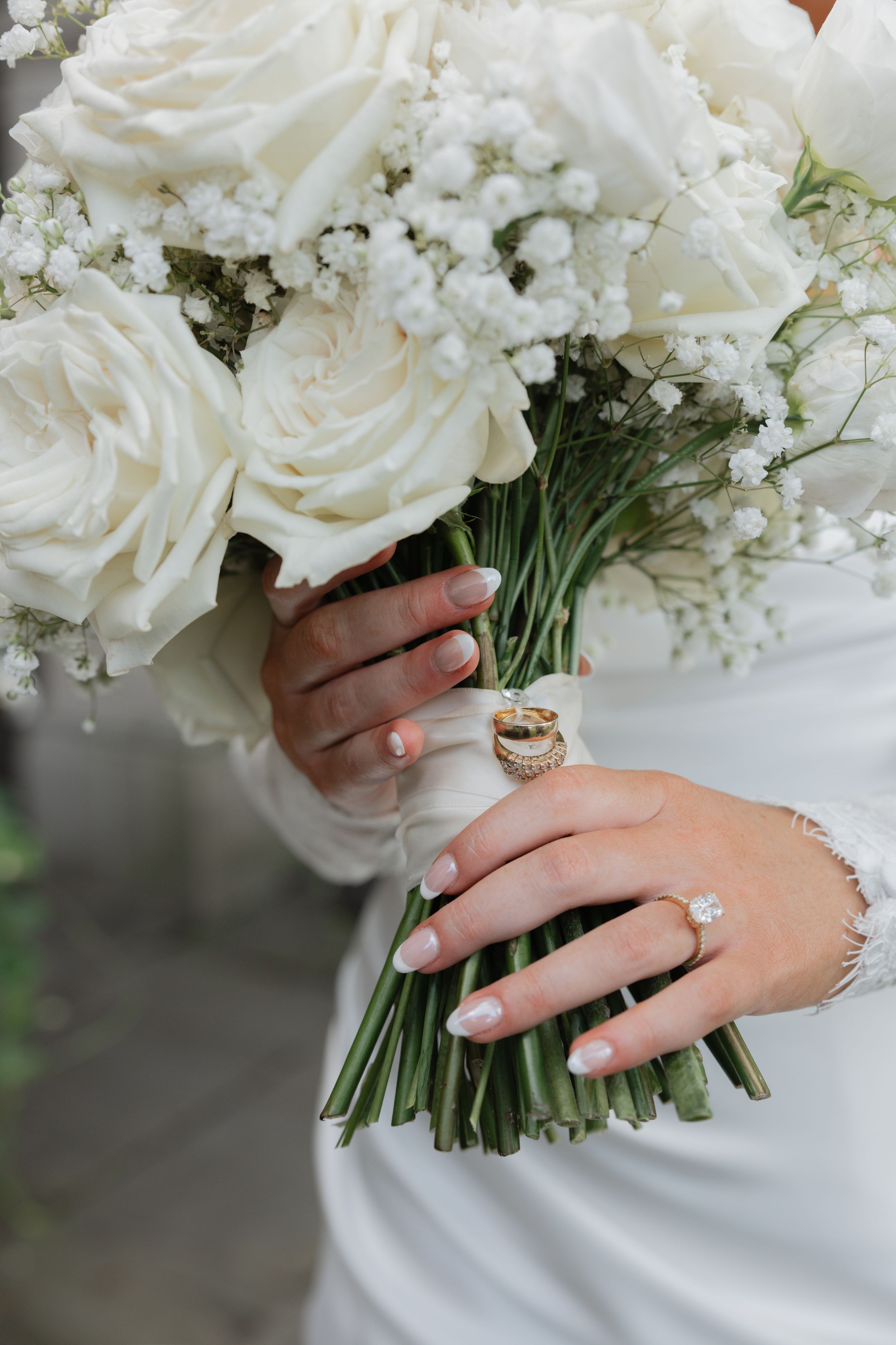 Close-up view of white wedding bouquet with roses and baby's breath being held by hands wearing elegant rings.