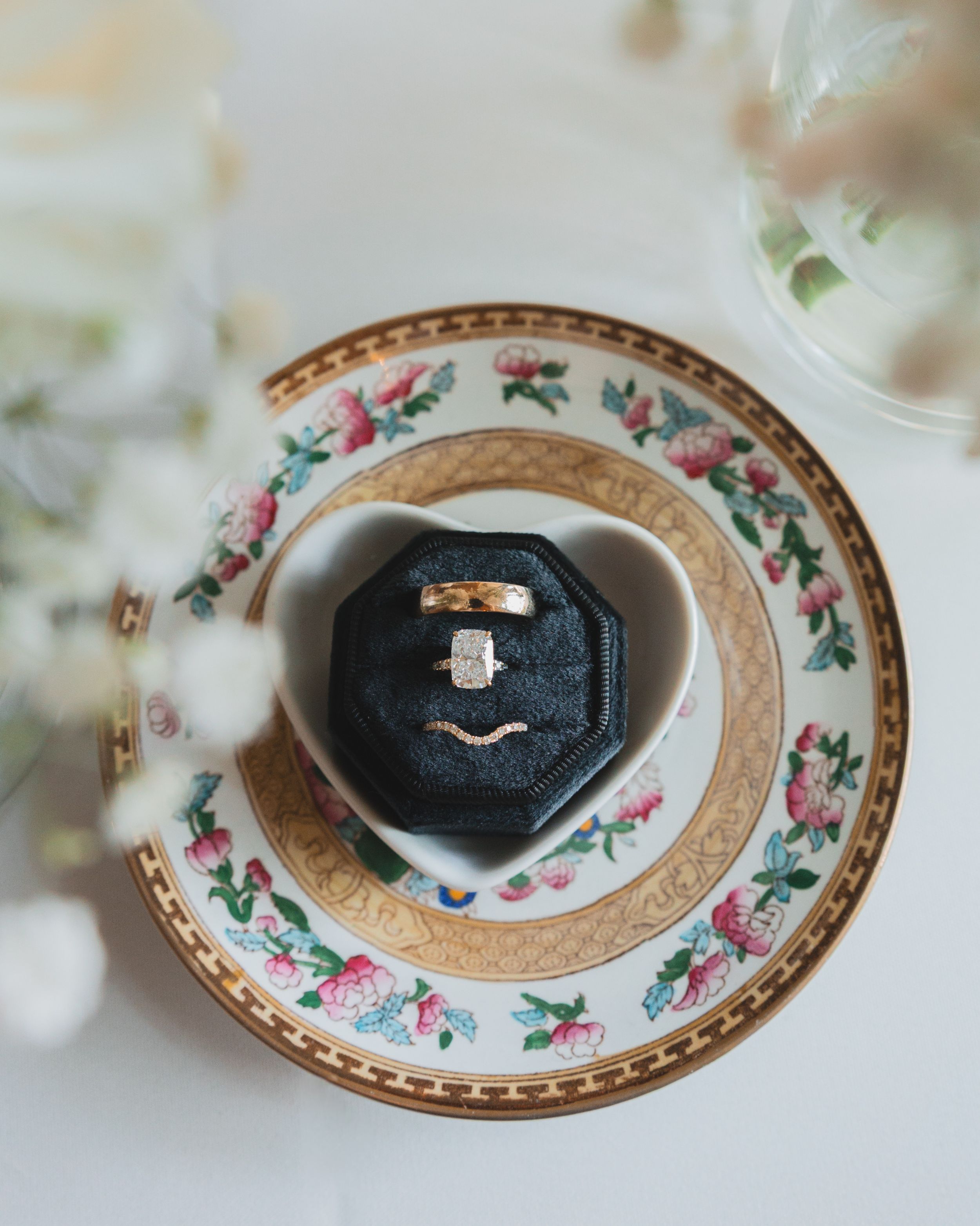 Wedding rings displayed on a vintage floral patterned china plate with gold trim and pink rose details.