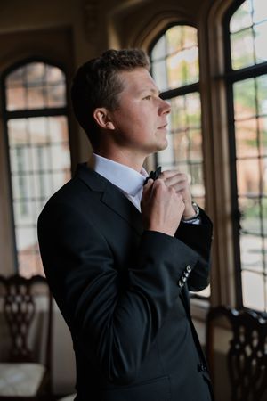 A person in a black suit stands thoughtfully by a window in a sequence of four similar poses.