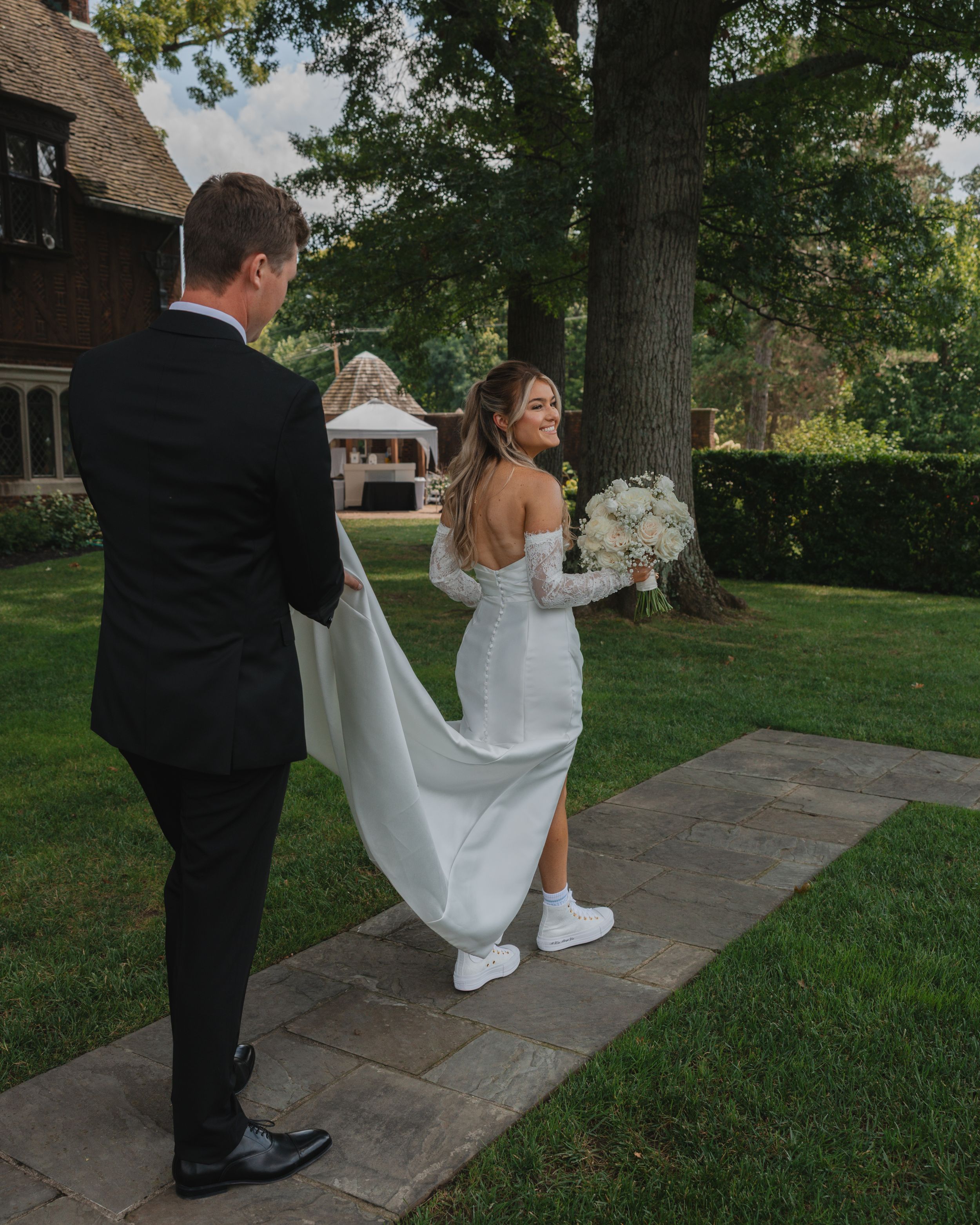 A couple walks along a stone path through a green lawn during an outdoor wedding celebration.