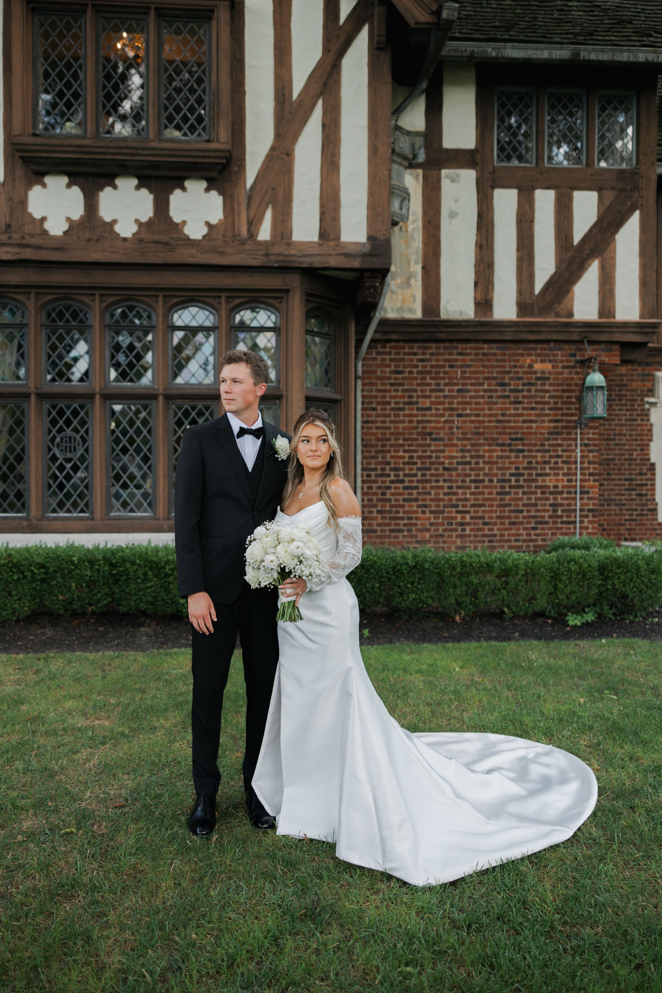 A couple in wedding attire poses outside a Tudor-style mansion with brick walls and decorative windows.