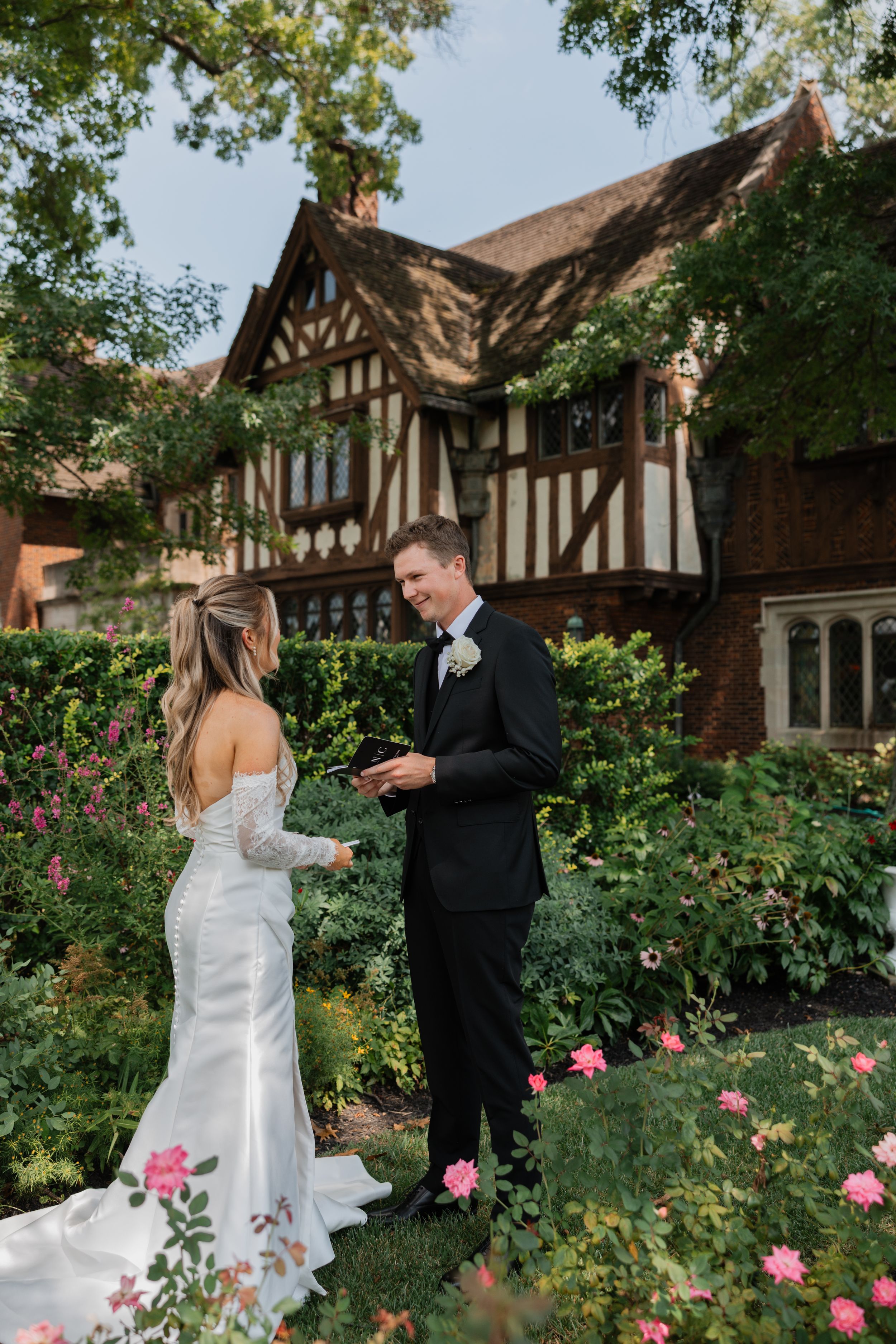 A couple shares a romantic moment in a garden with blooming pink roses and Tudor-style architecture in the background.