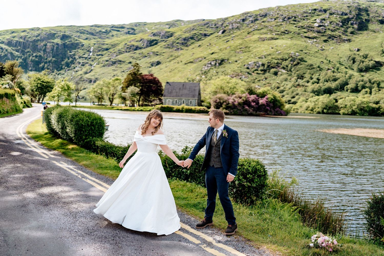 A couple in wedding attire celebrates their special day along a scenic riverside road with green hills in the background.
