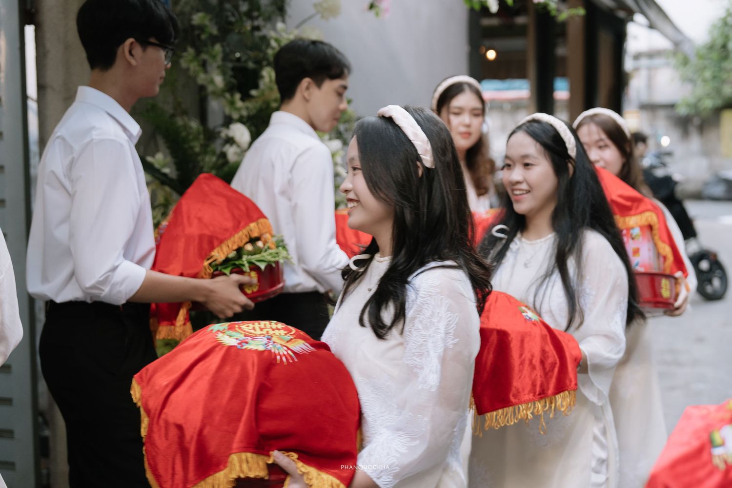 Smiling bridesmaids in white ao dai carrying red ceremonial gift trays at a traditional Vietnamese wedding.