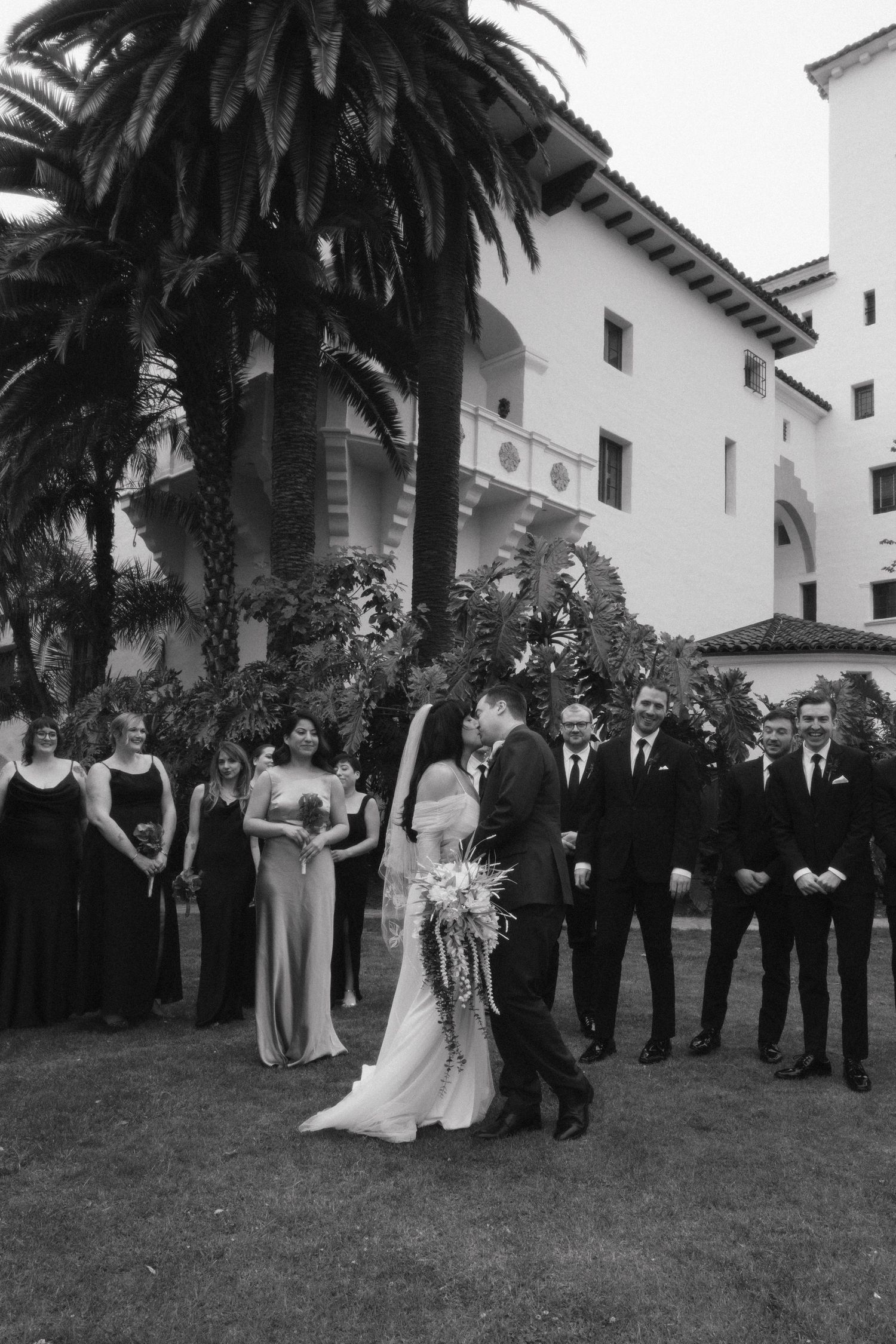 Black and white wedding party formal photo lineup outdoors with palm trees and Spanish colonial building in background.