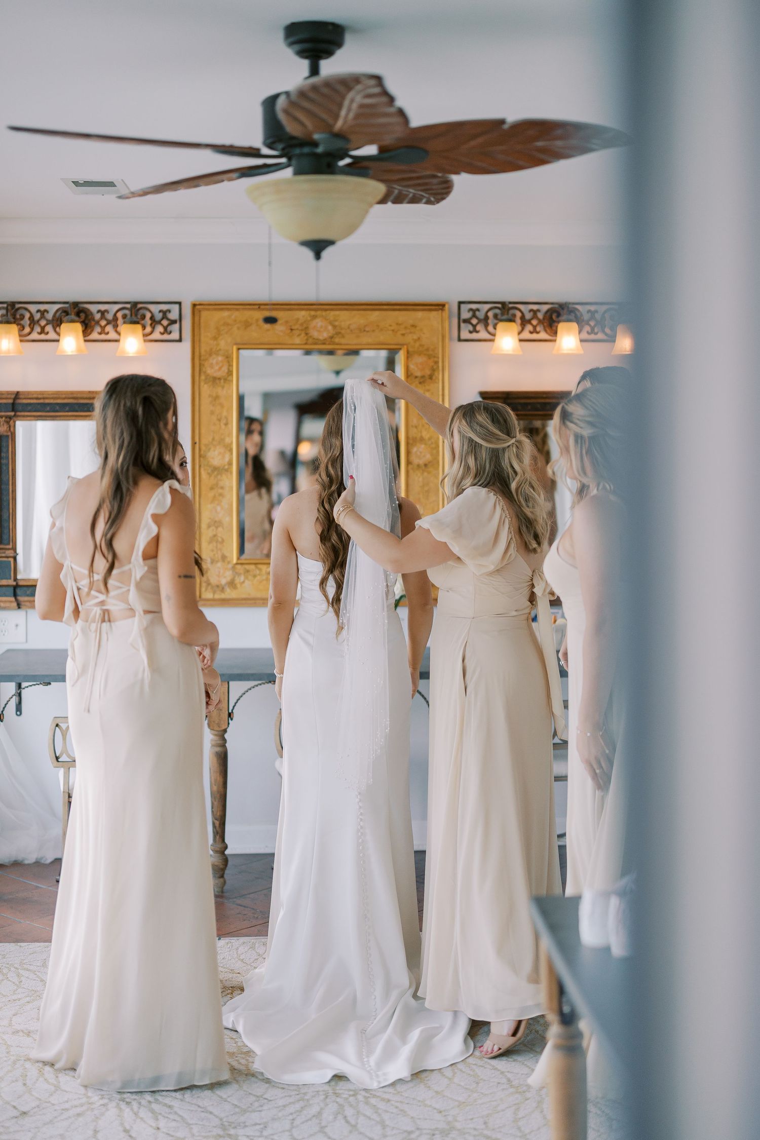 Row of bridesmaids in flowing white dresses standing together from behind in a sunlit room.