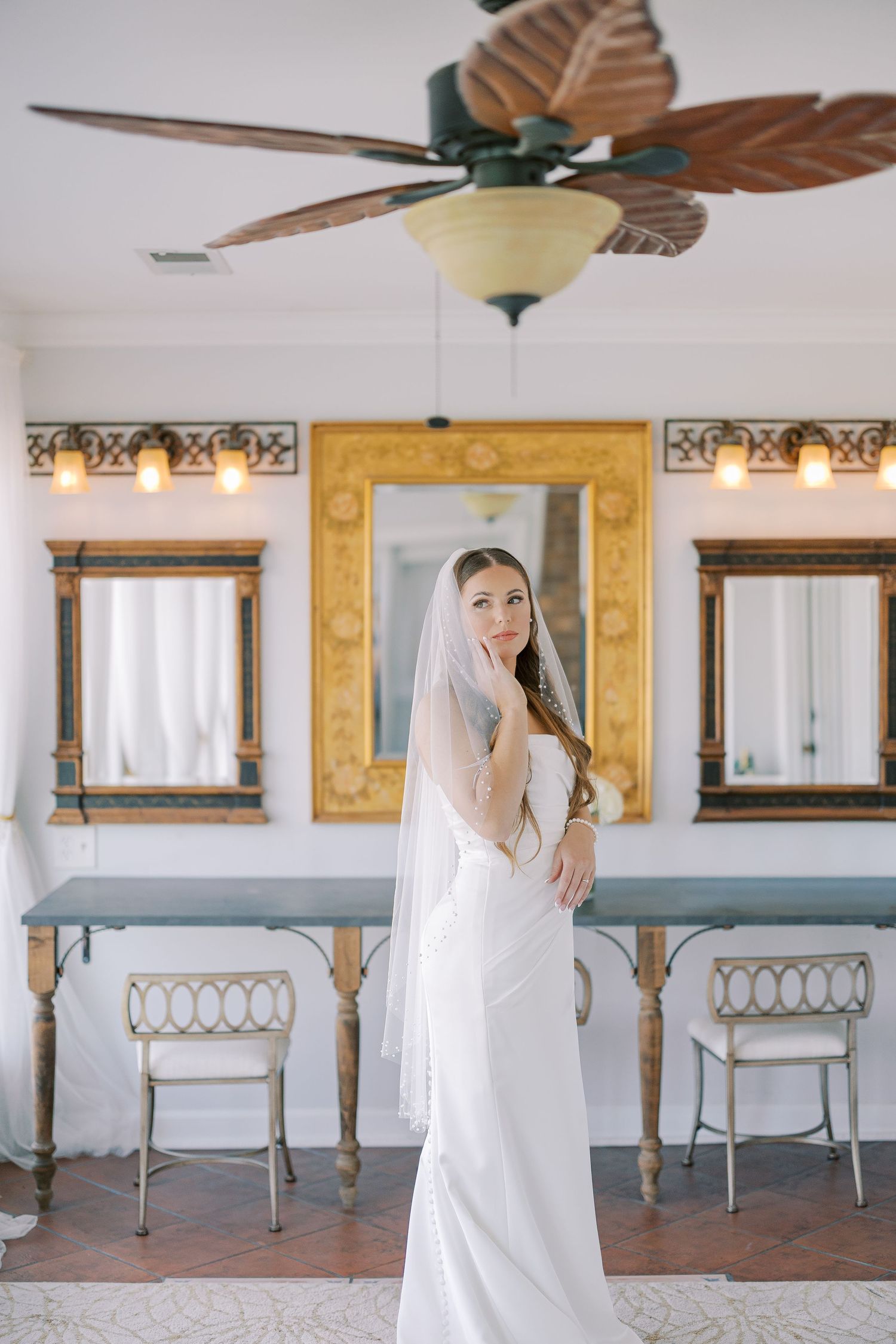 Elegant bridal portrait in white gown with flowing veil against ornate interior with gold accents and vintage chairs.