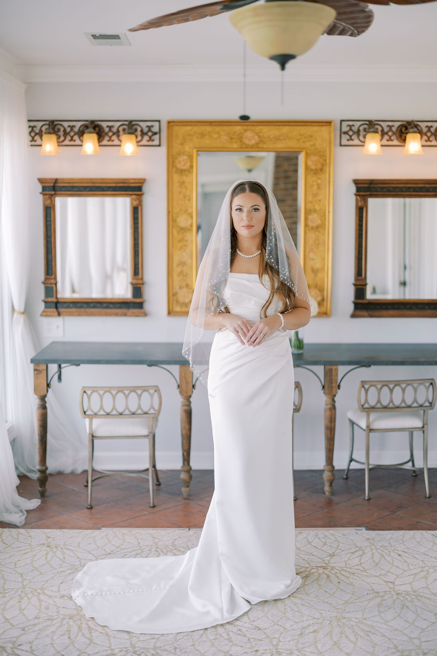 Bride in classic white dress and cathedral veil poses in historic venue with decorative gold trim and white chairs.