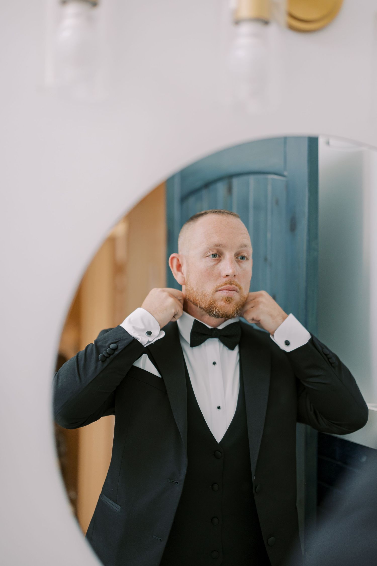 A groom in a black tuxedo and bow tie adjusts his collar while getting ready for a formal occasion.