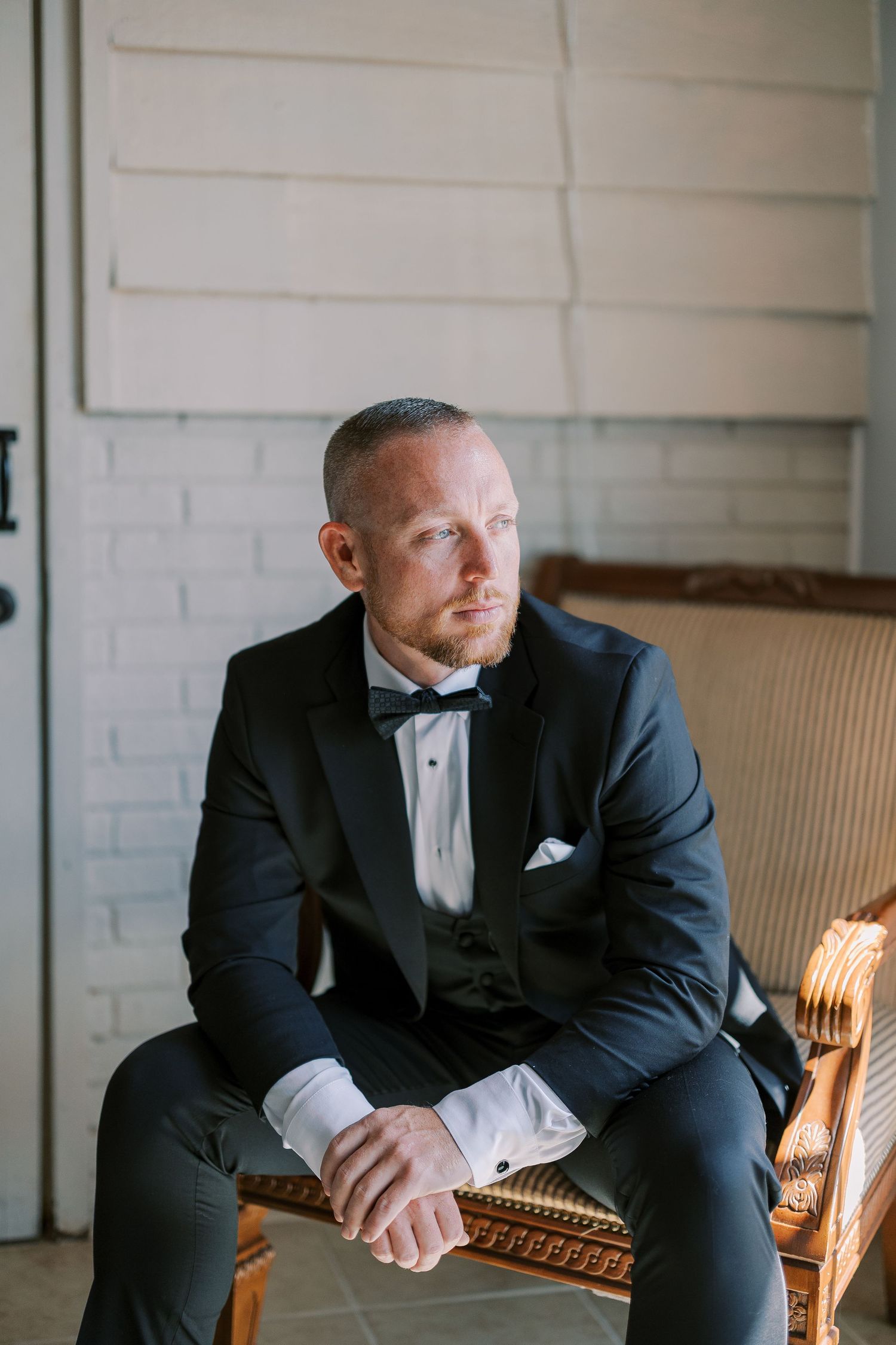 A groom in a classic black tuxedo and bow tie sits thoughtfully on an elegant beige couch in a brick-walled room.