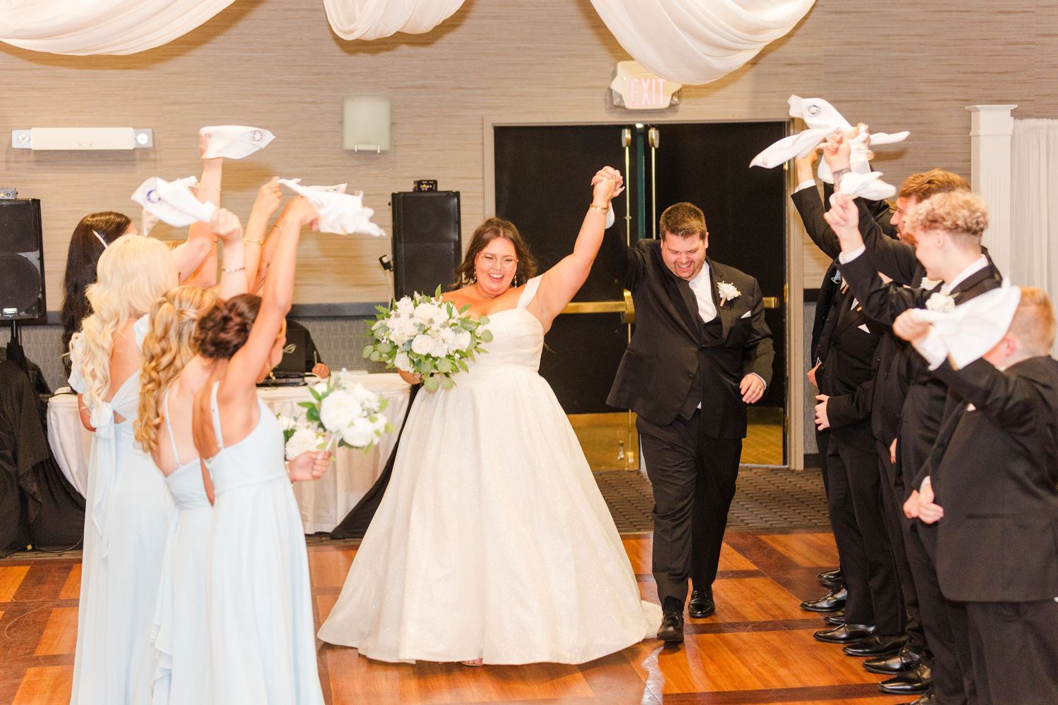 Wedding party celebrates with joy and excitement on the dance floor as newlyweds raise their arms triumphantly.