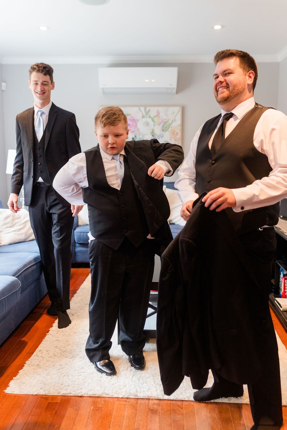 Groomsmen getting ready for wedding by helping each other adjust their formal attire in a bright room.