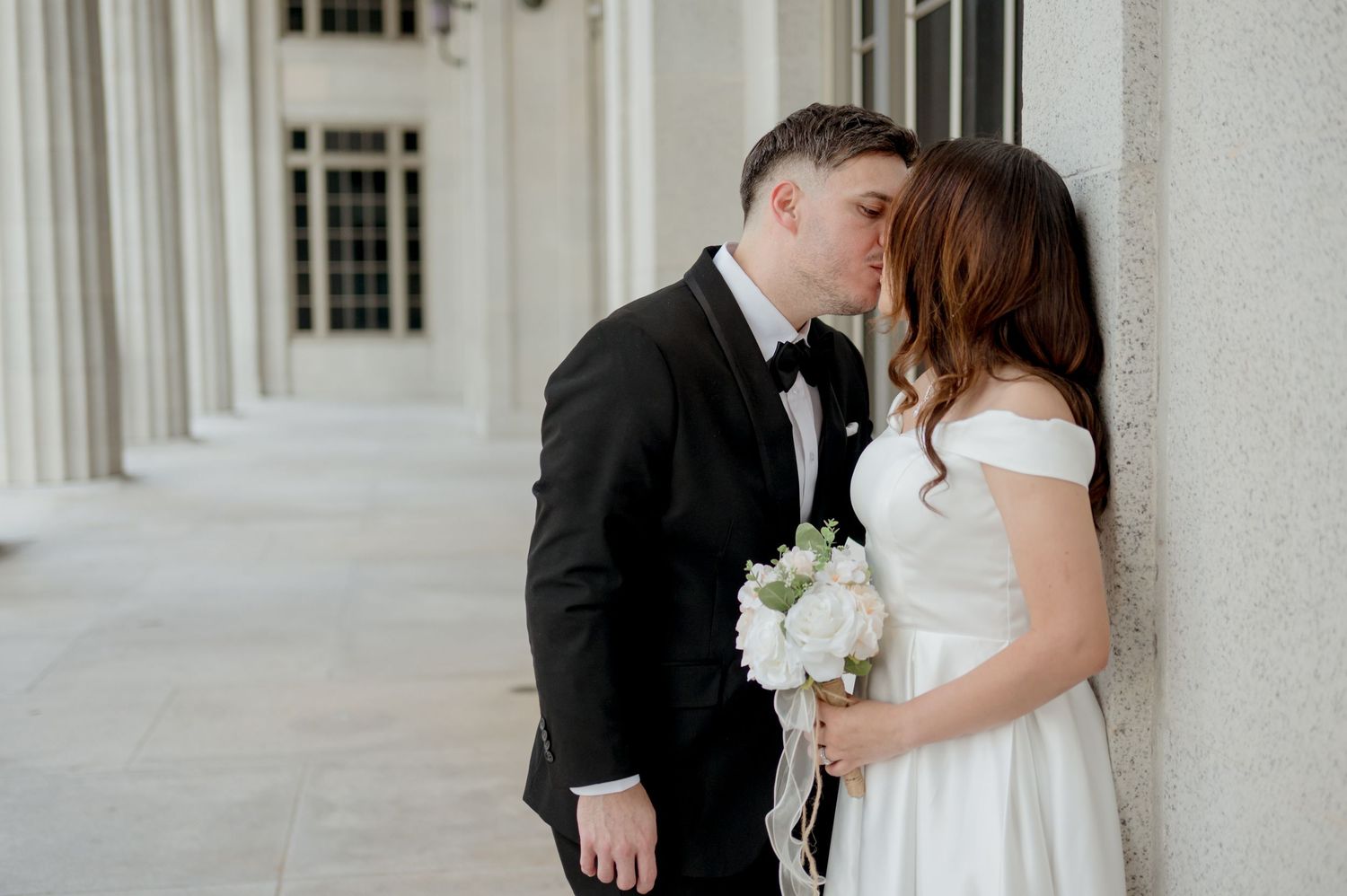 Couple sharing romantic kisses during outdoor wedding photo session against white column backdrop.