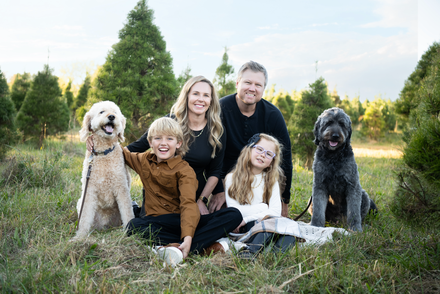 Family and their pets pose together outdoors among evergreen trees on a sunny day.