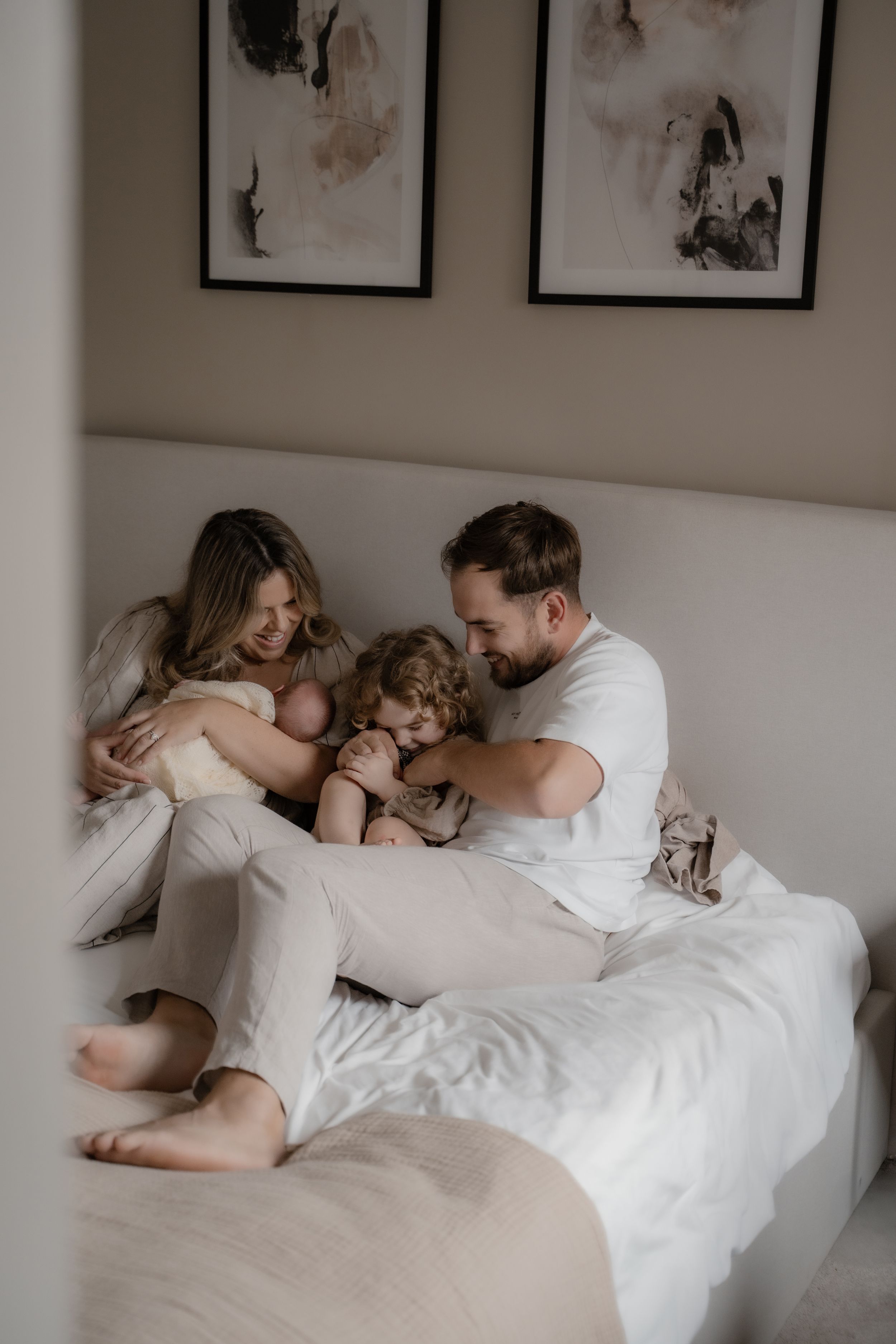 A family relaxes together on a white bed in a cozy bedroom with framed artwork on the wall.