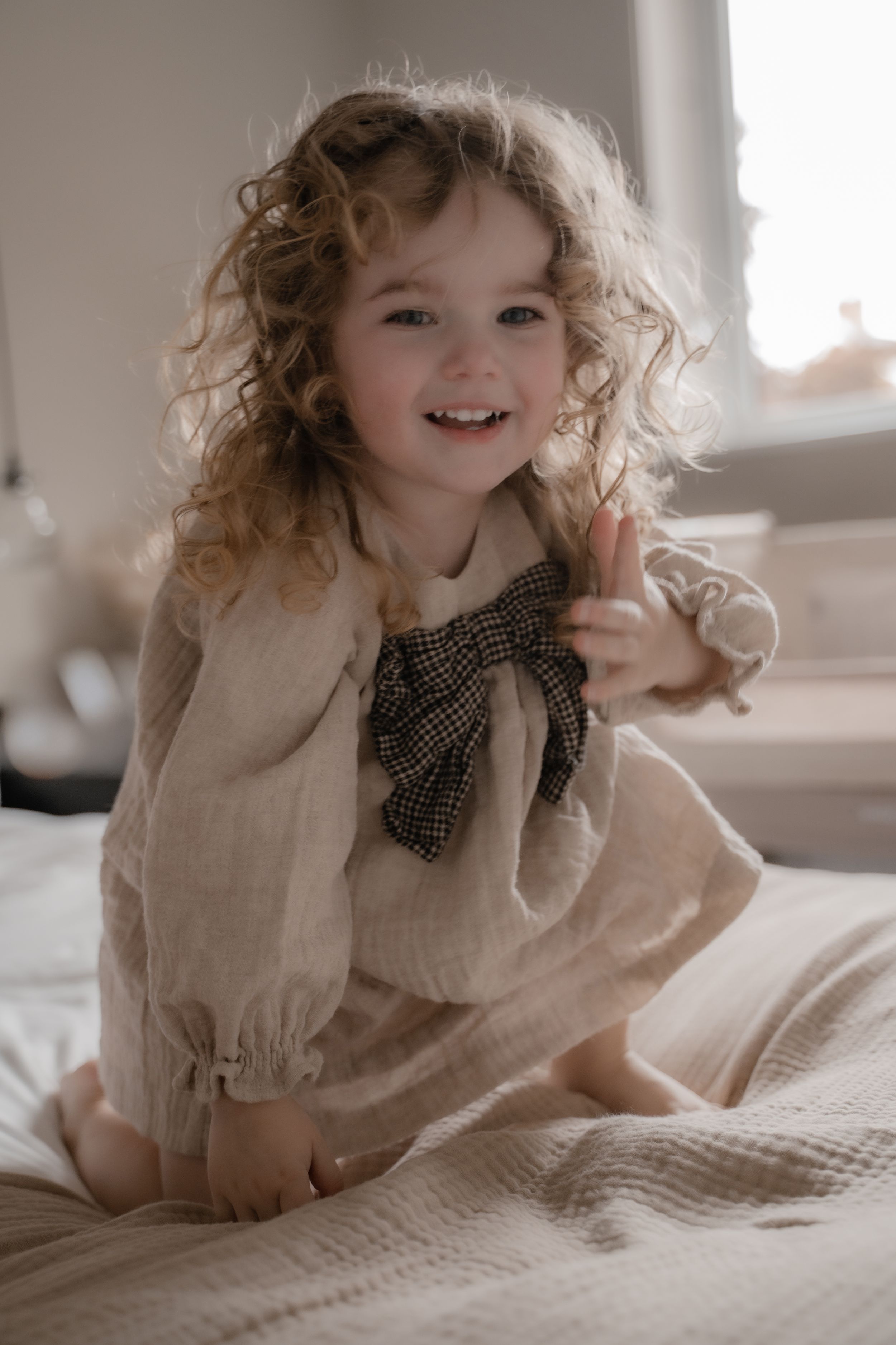 A child with curly hair wearing a beige linen dress with a plaid bow sits happily on a bed.