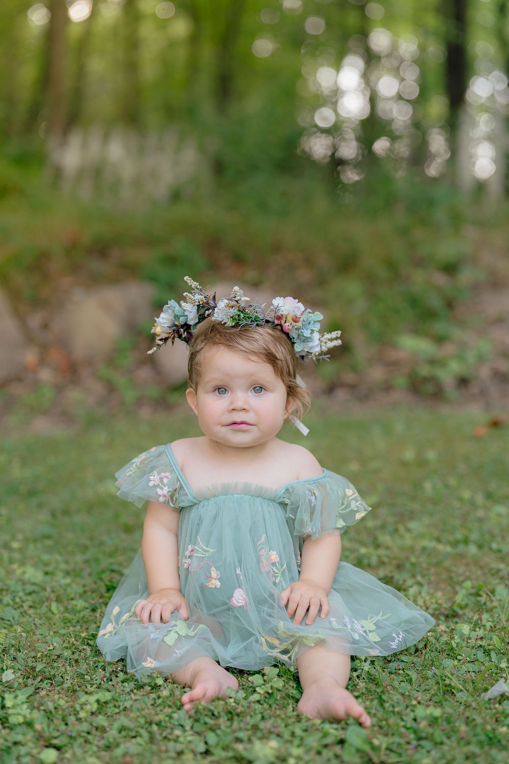 A baby wearing a mint green tulle dress and floral crown sitting on grass in a dreamy outdoor setting at One Year Milestone Session in Akron