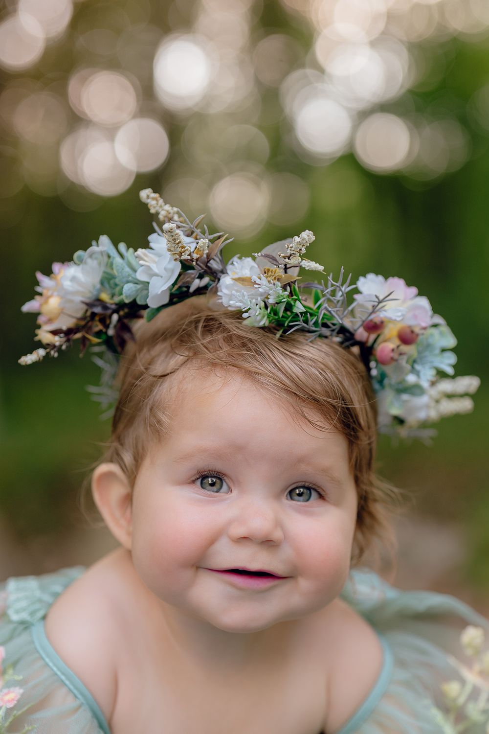 A child wearing a mint green dress and floral crown smiles at the camera with bokeh background effect in Akron, Ohio One Year Photo Session