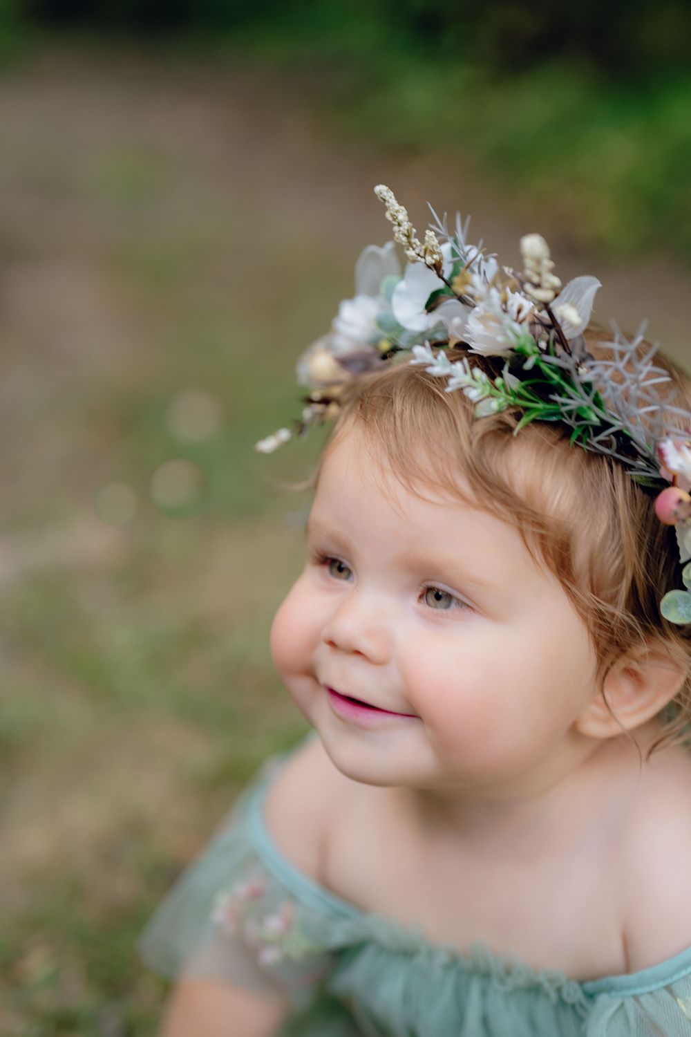 A child wearing a delicate flower crown and mint green dress looks up with a gentle smile in an outdoor setting in Akron, Ohio 1 Year Photo