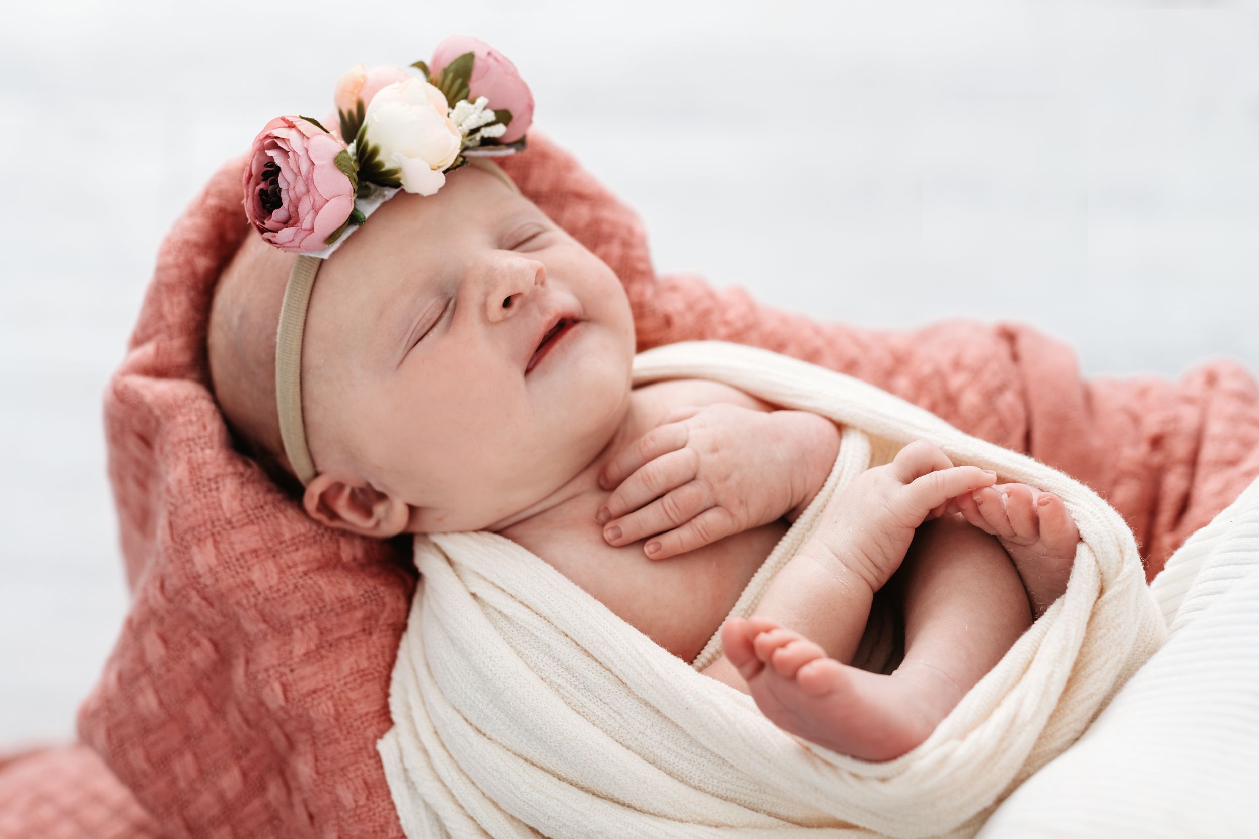 A sleeping newborn baby wearing a floral headband wrapped in white fabric against coral pink backdrop.