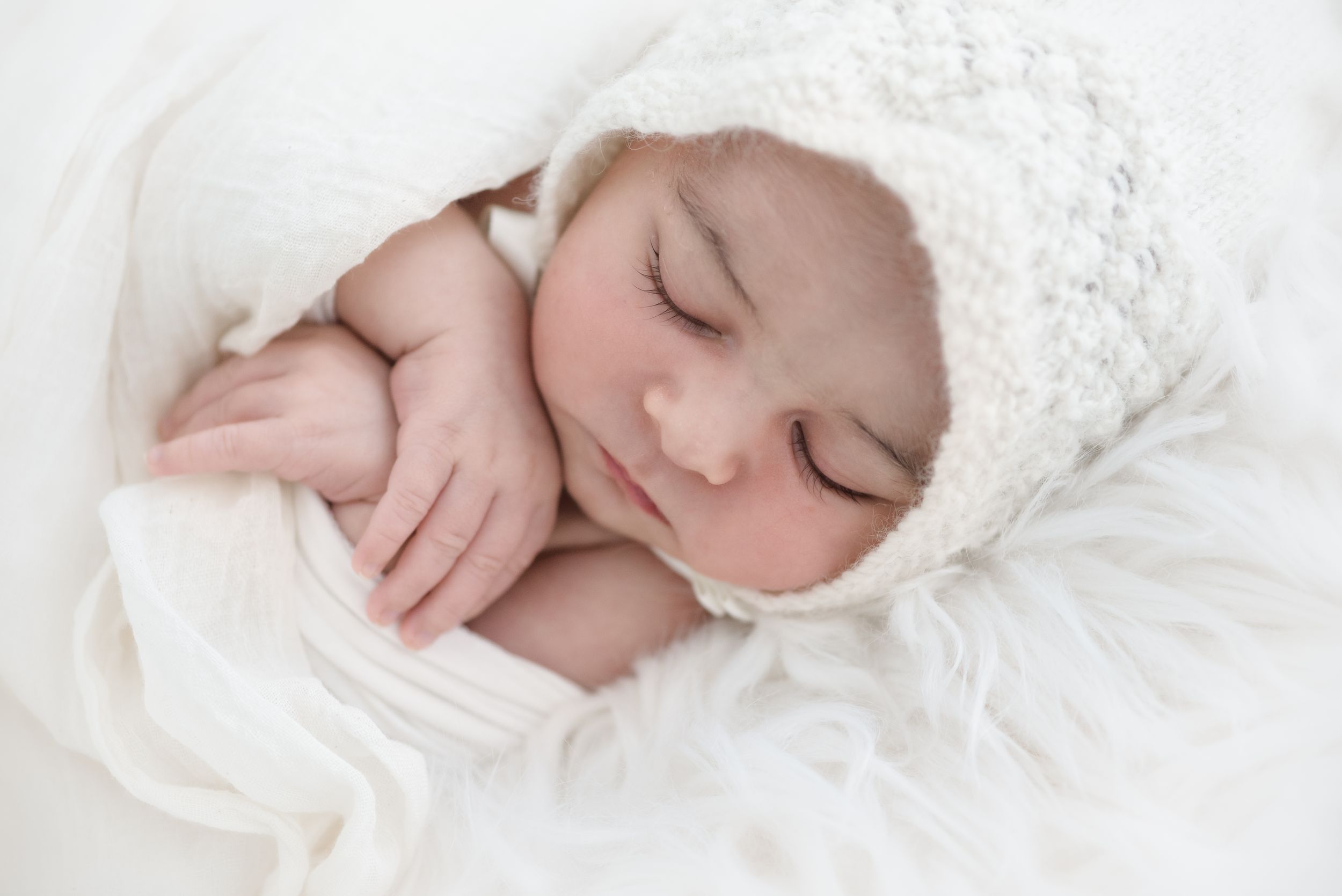 Sleeping newborn wearing white lace bonnet wrapped in soft white fabric.