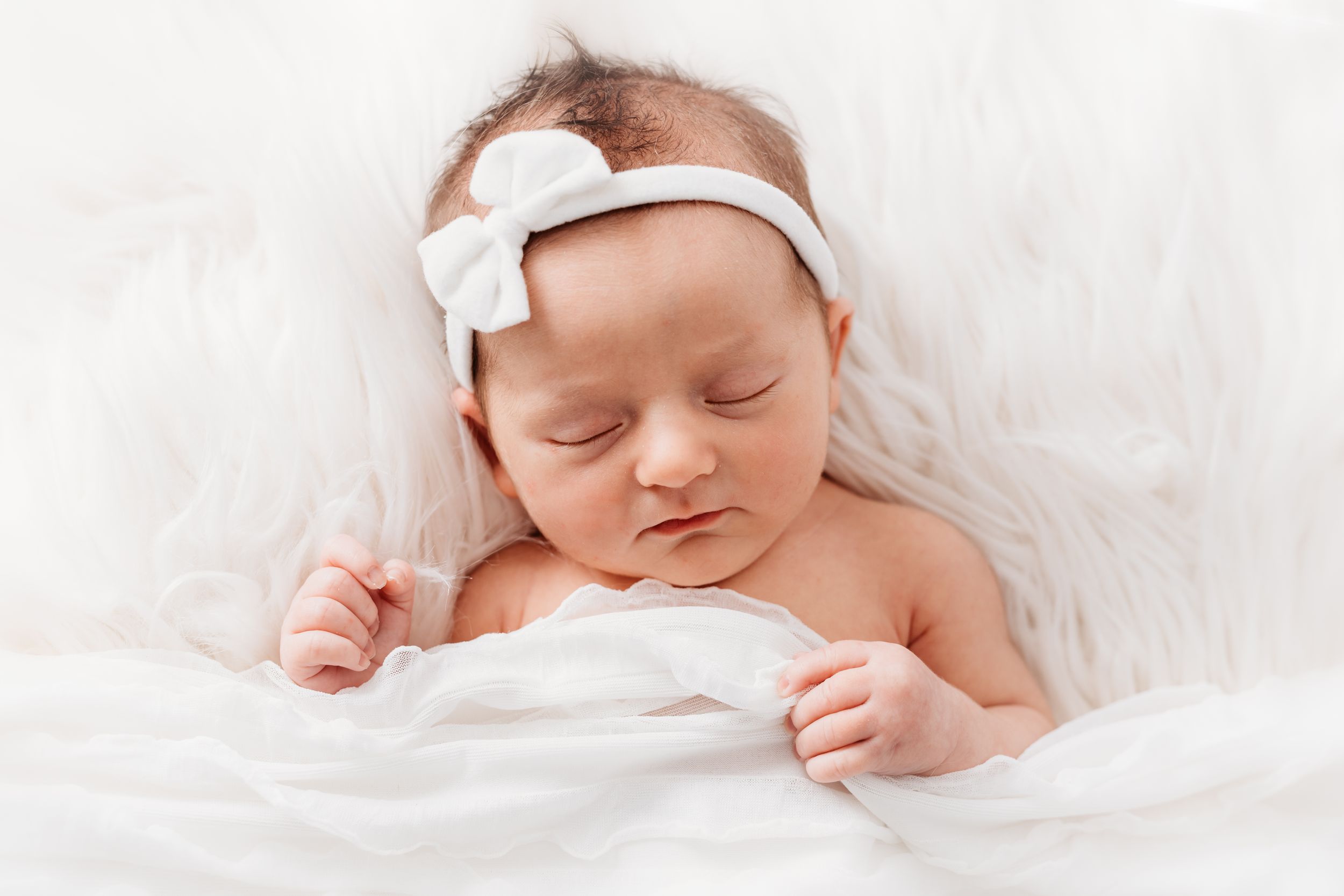 Peaceful sleeping newborn baby wearing white headband bow resting on soft white blanket.