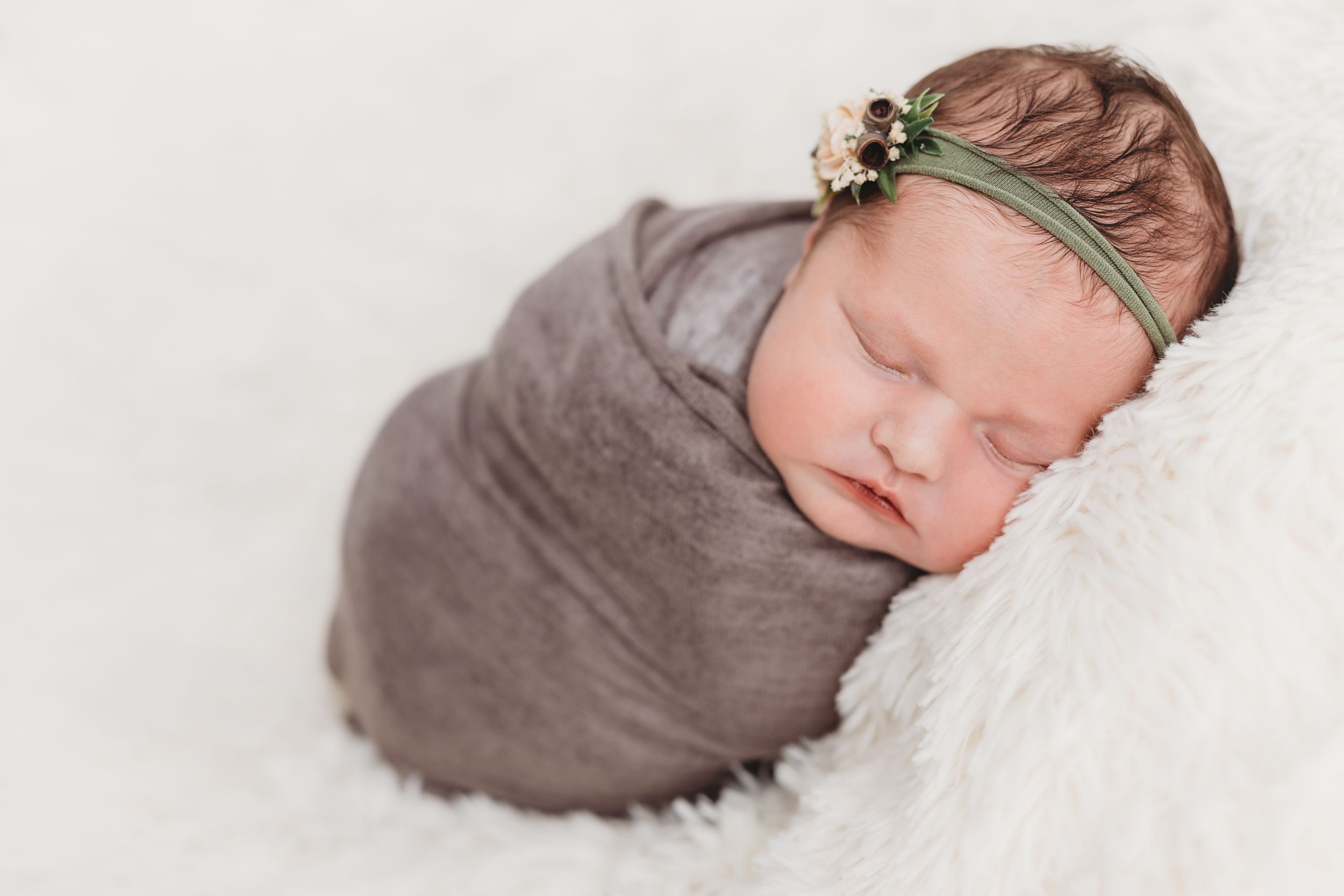 Sleeping newborn wrapped in grey swaddle wearing delicate floral headband on white fur backdrop.