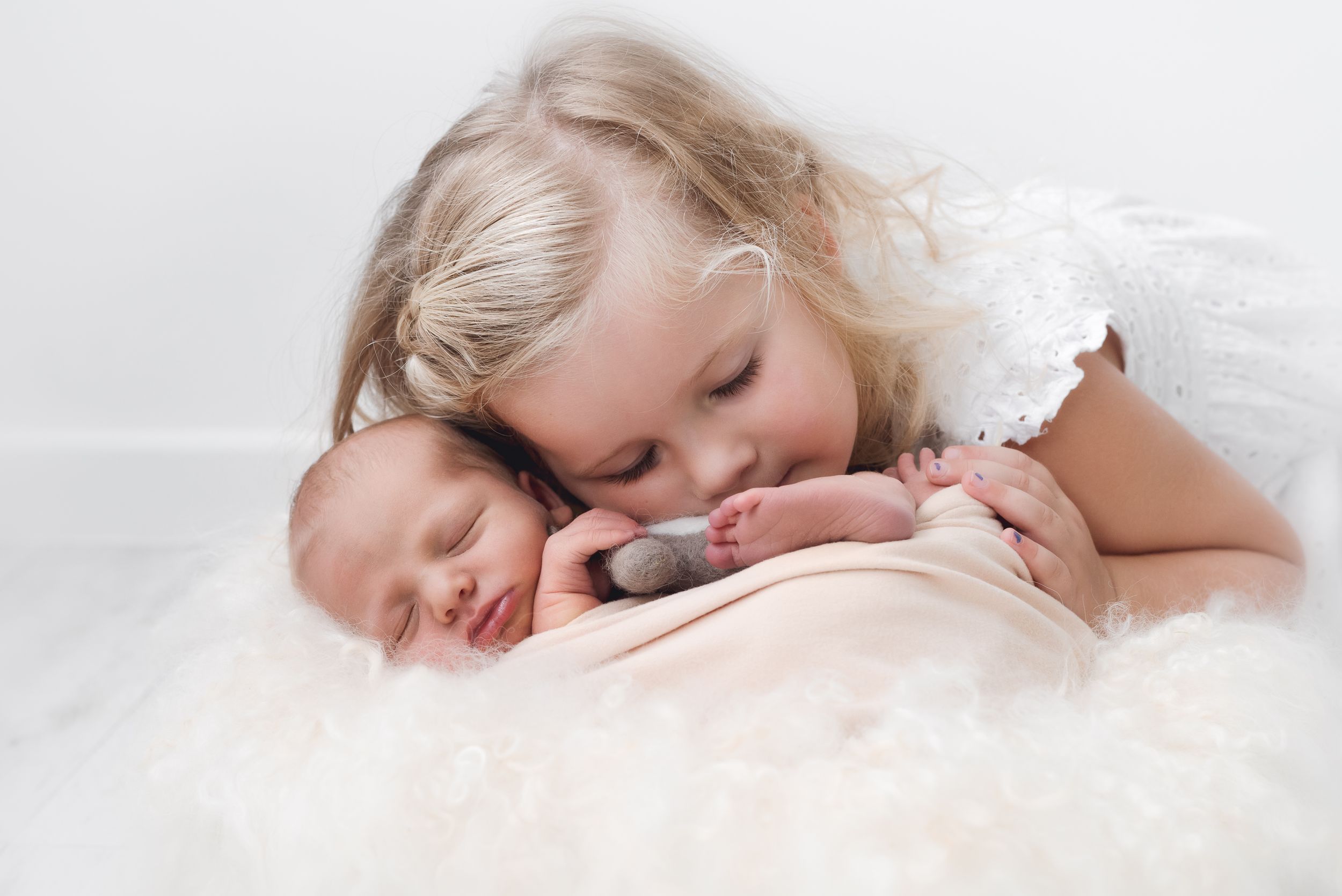 Soft portrait of sibling gently embracing newborn baby on white fabric.