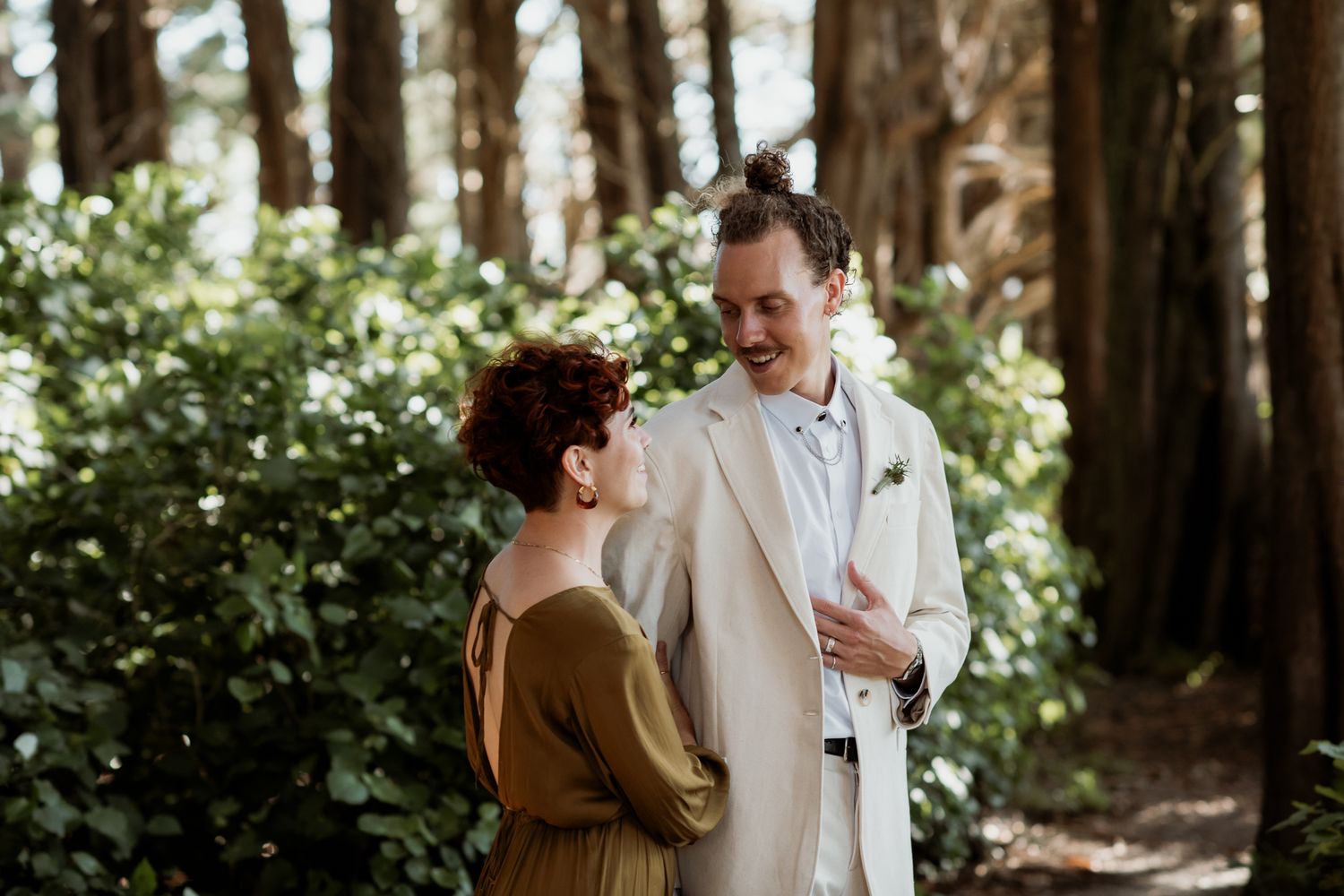 Married couple standing together in a romantic forest setting in Wairarapa with sunlight streaming through tall trees by August & Vine