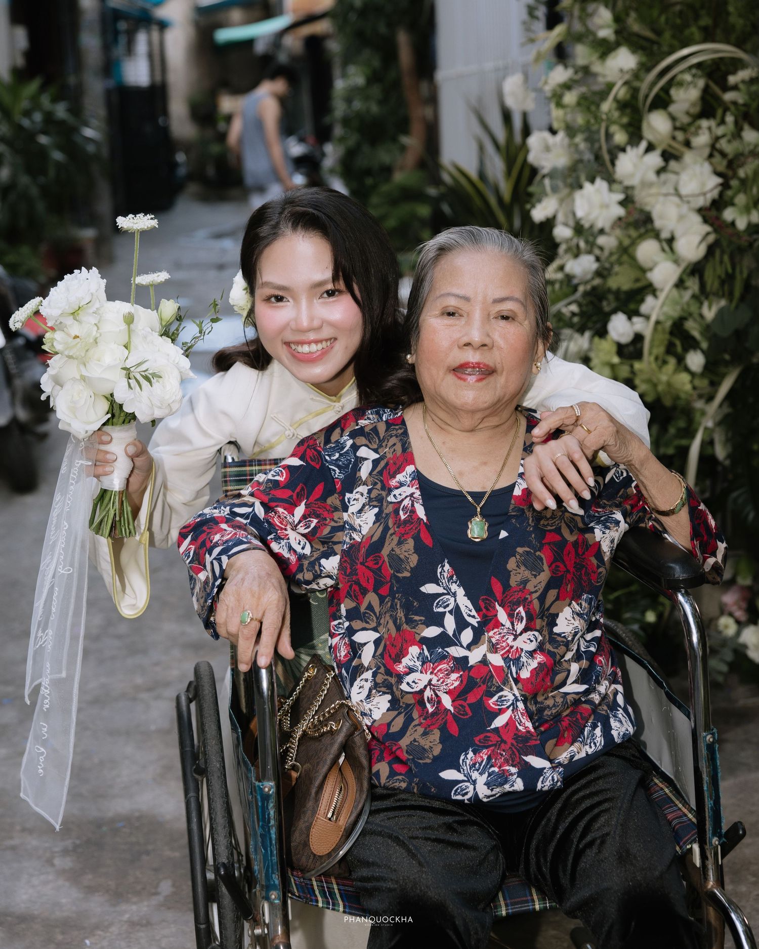 Smiling bride in white ao dai holds white bouquet posing with elderly woman in wheelchair.