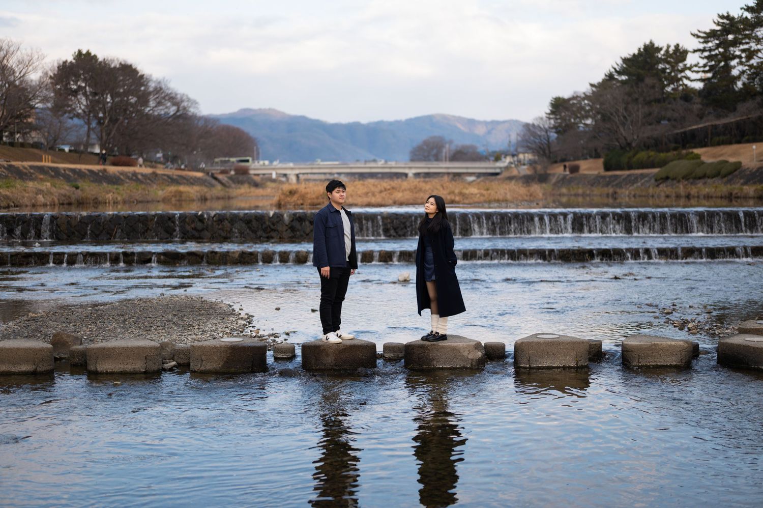 Couple standing on stepping stones across a shallow river with waterfall and mountains behind.