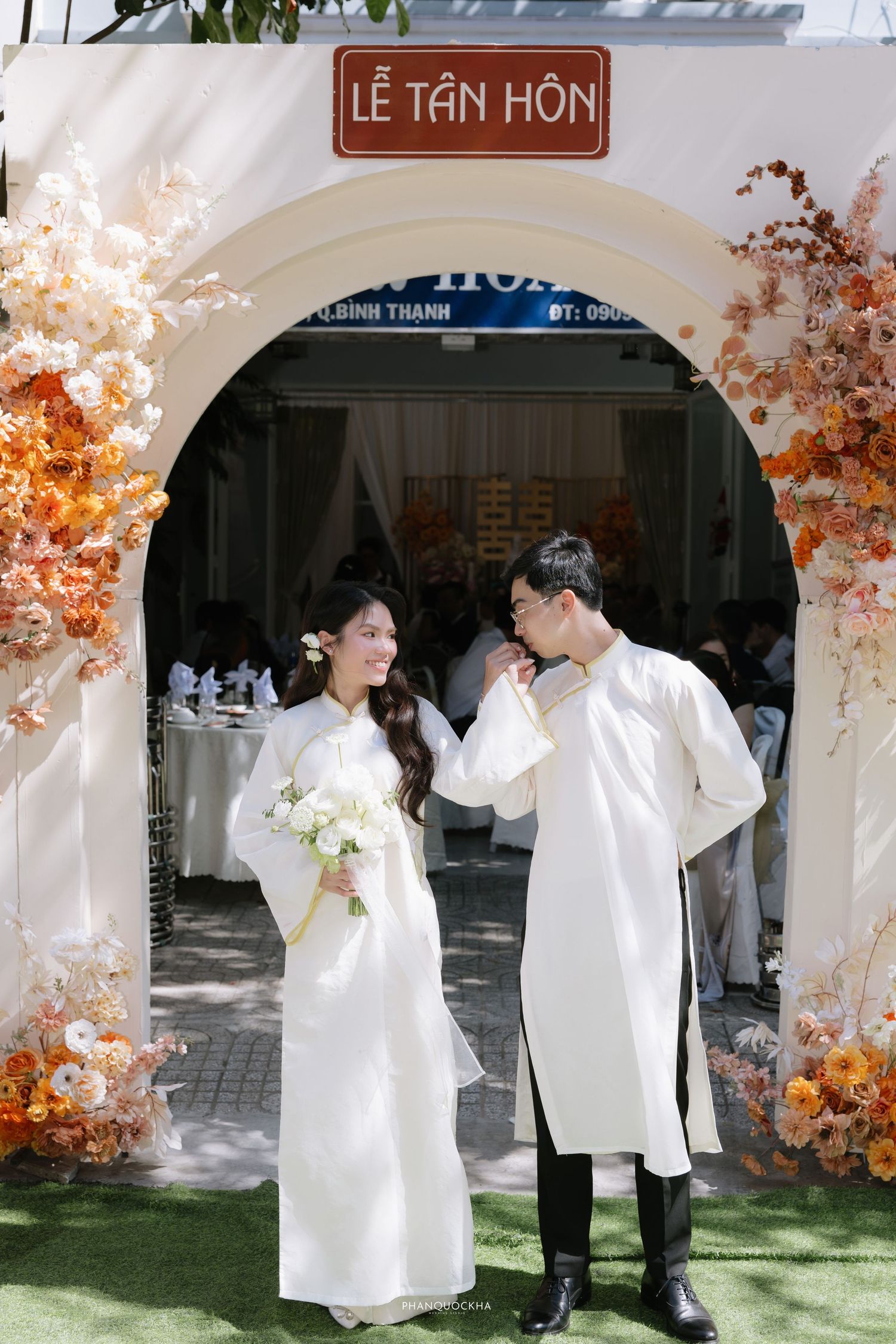 Vietnamese bride and groom in white ao dai standing under Le Tan Hon arch with orange floral decor.