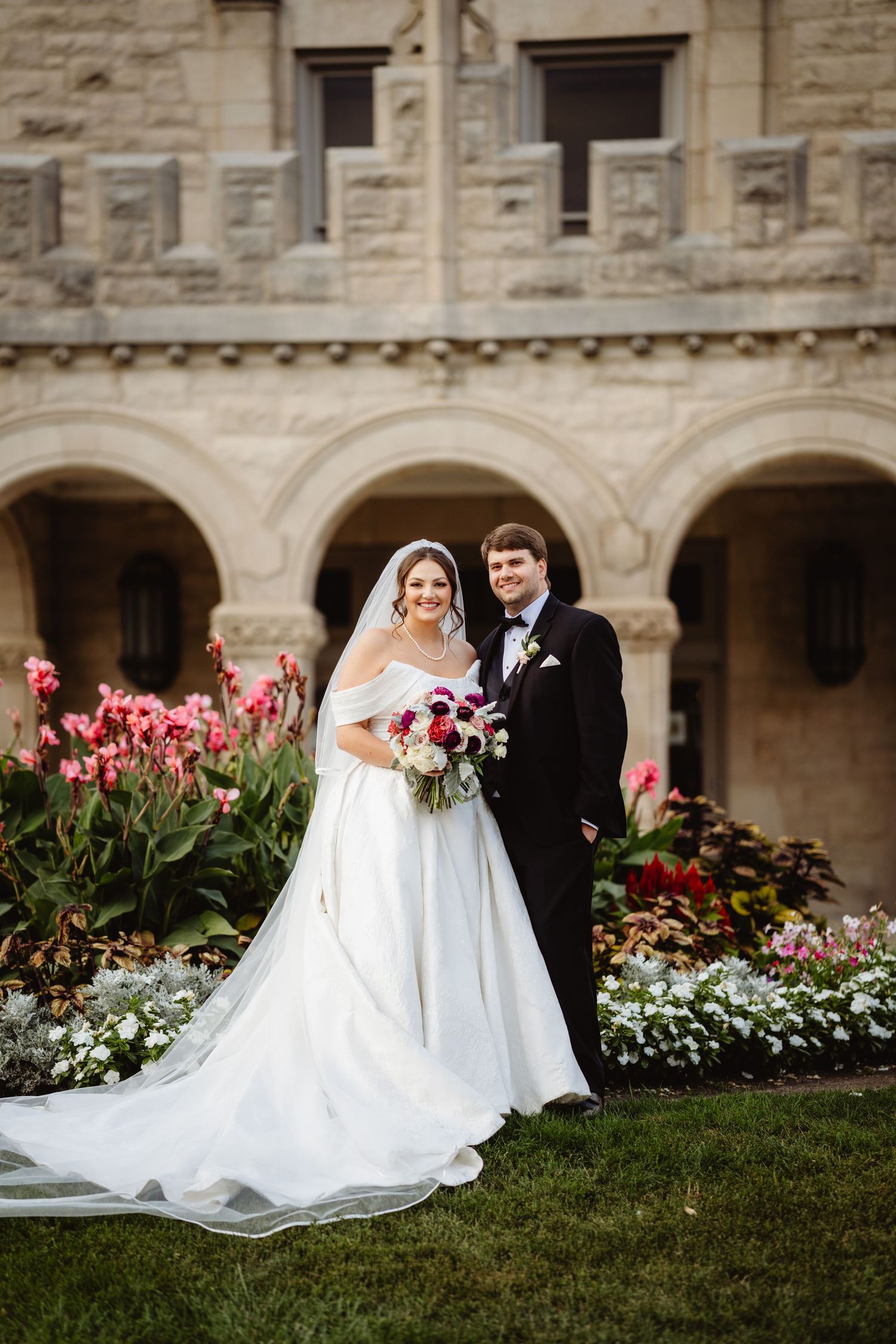 Newlywed couple standing near blooming pink tulips and white flowers against a stone building exterior.