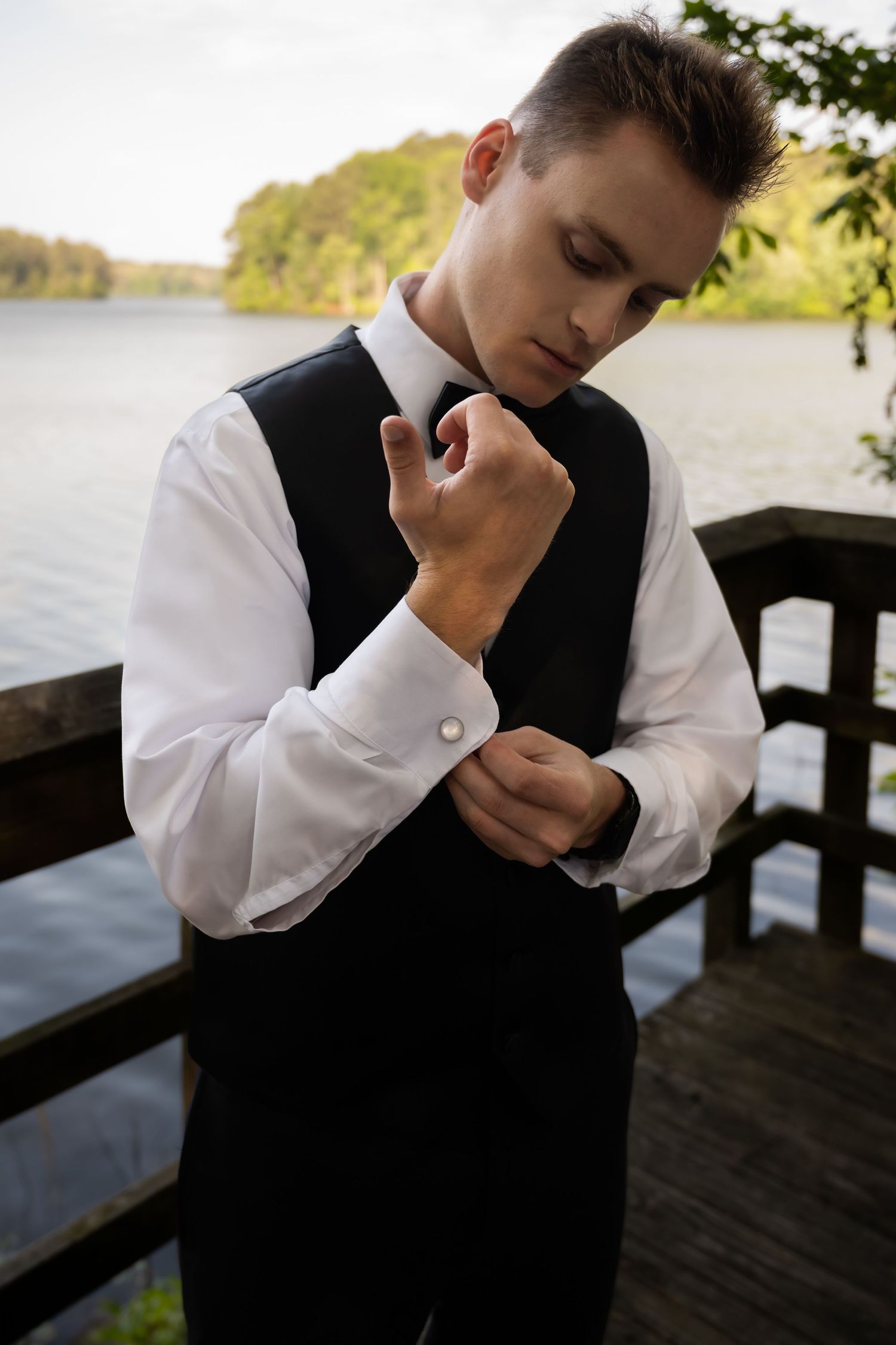 Groom standing on a wooden deck by a lake adjusting their shirt cuffs.