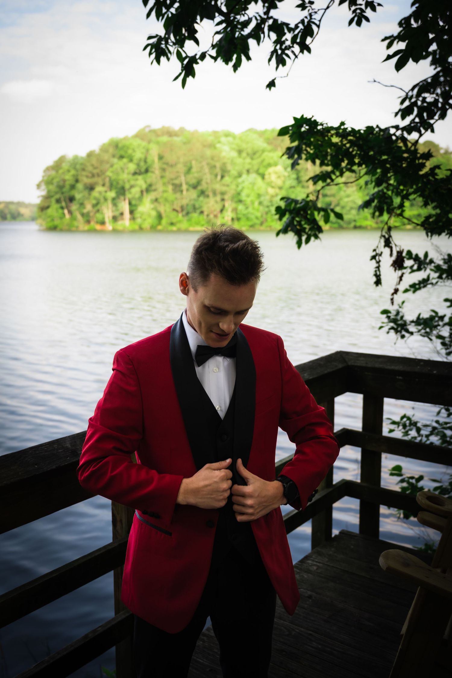 Groom in red tuxedo jacket adjusts their outfit on dock overlooking lake.