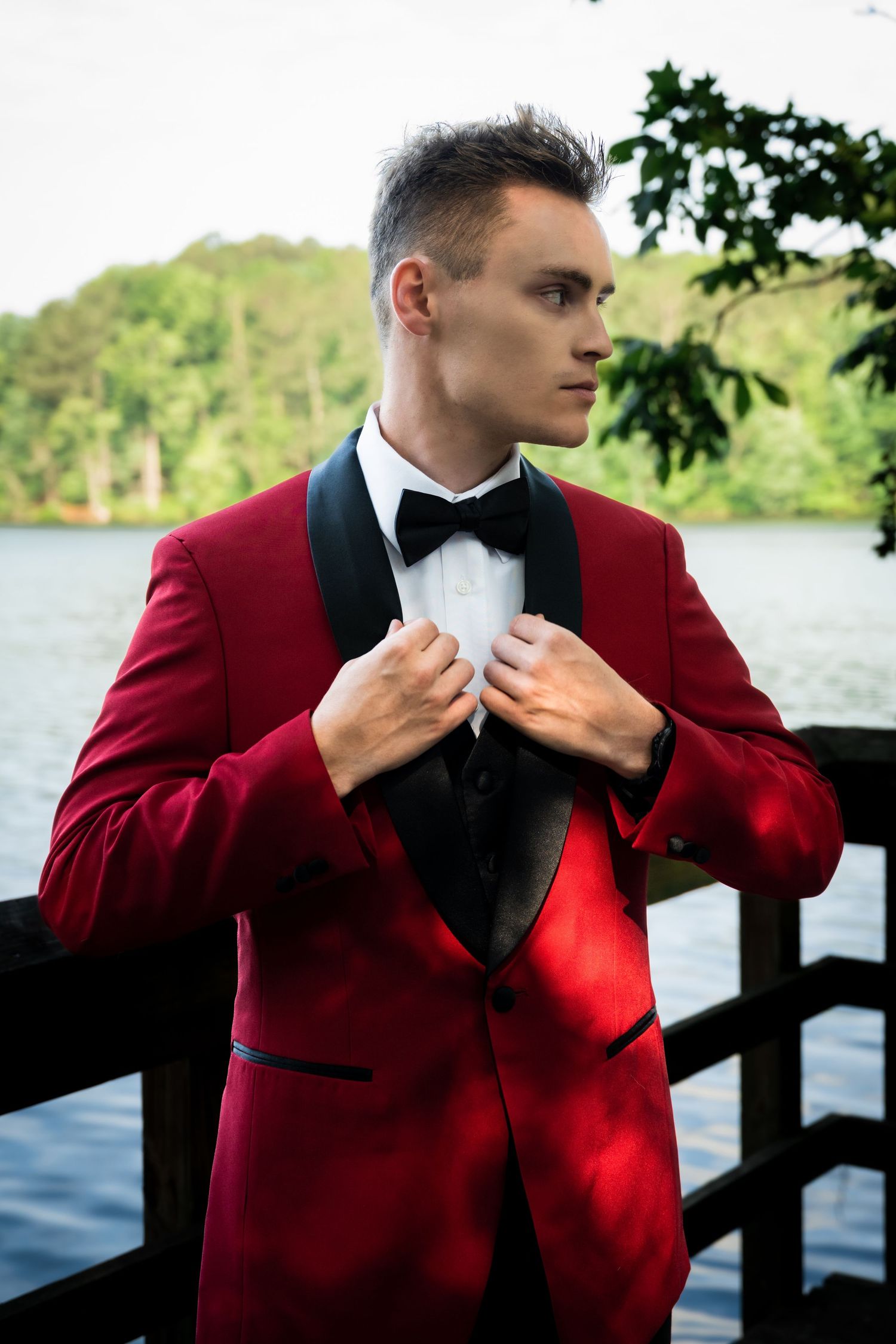Groom in burgundy tuxedo jacket poses against lakeside backdrop.