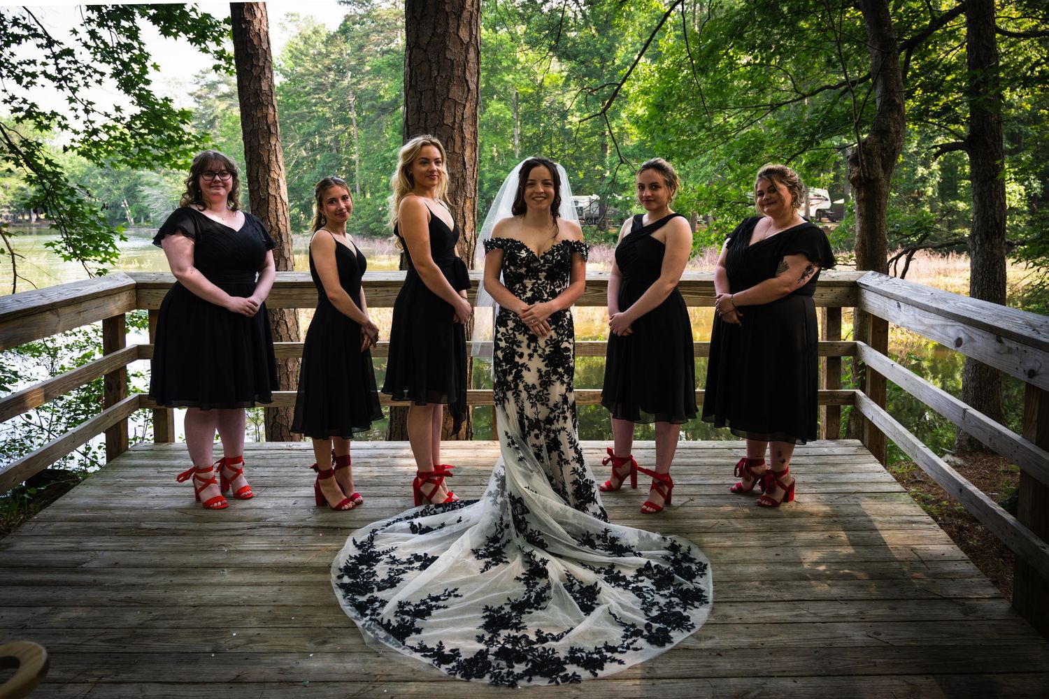 Bridal party in black dresses seated on wooden deck stairs with lake view.