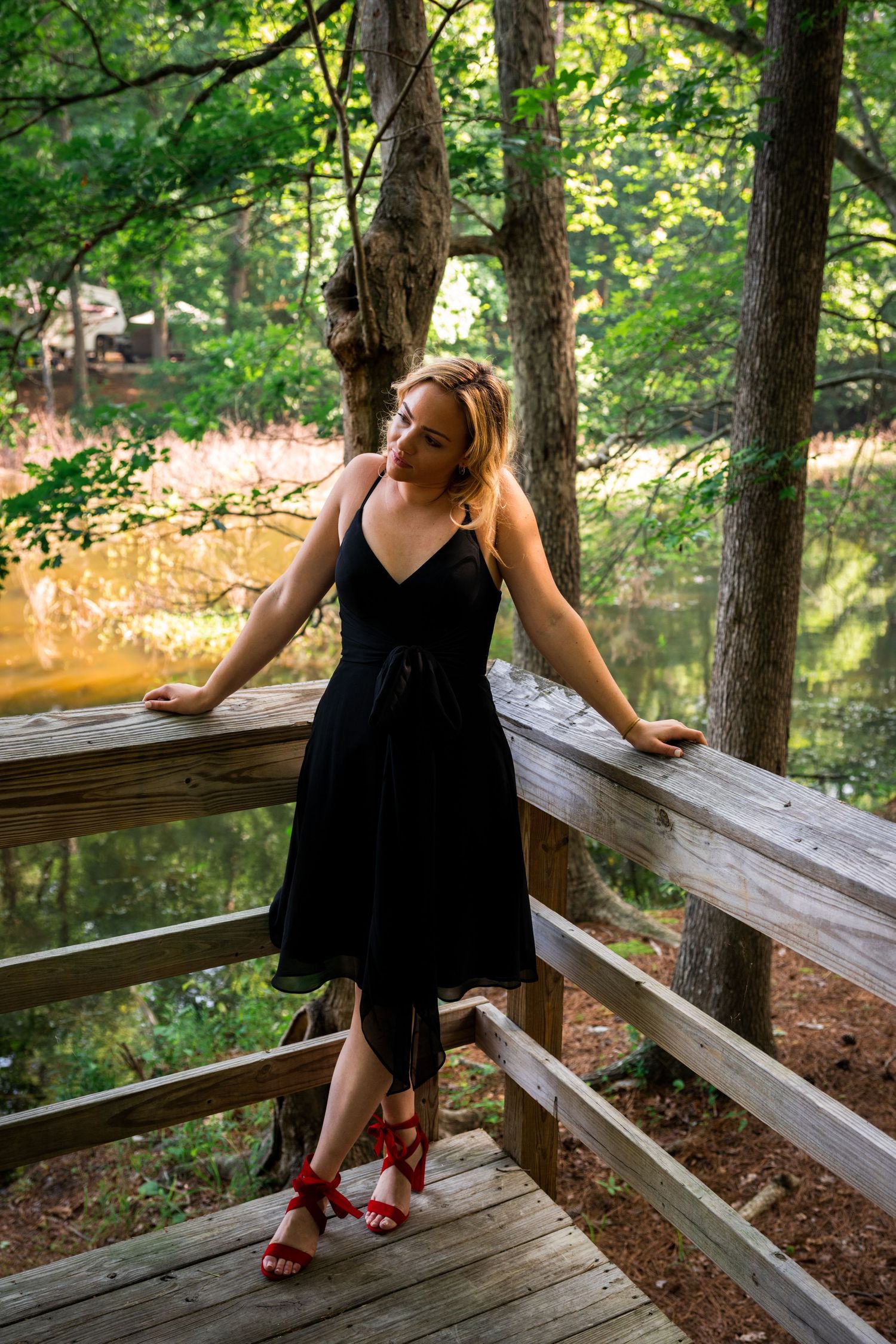 Woman in black dress poses casually on wooden deck railing near water.