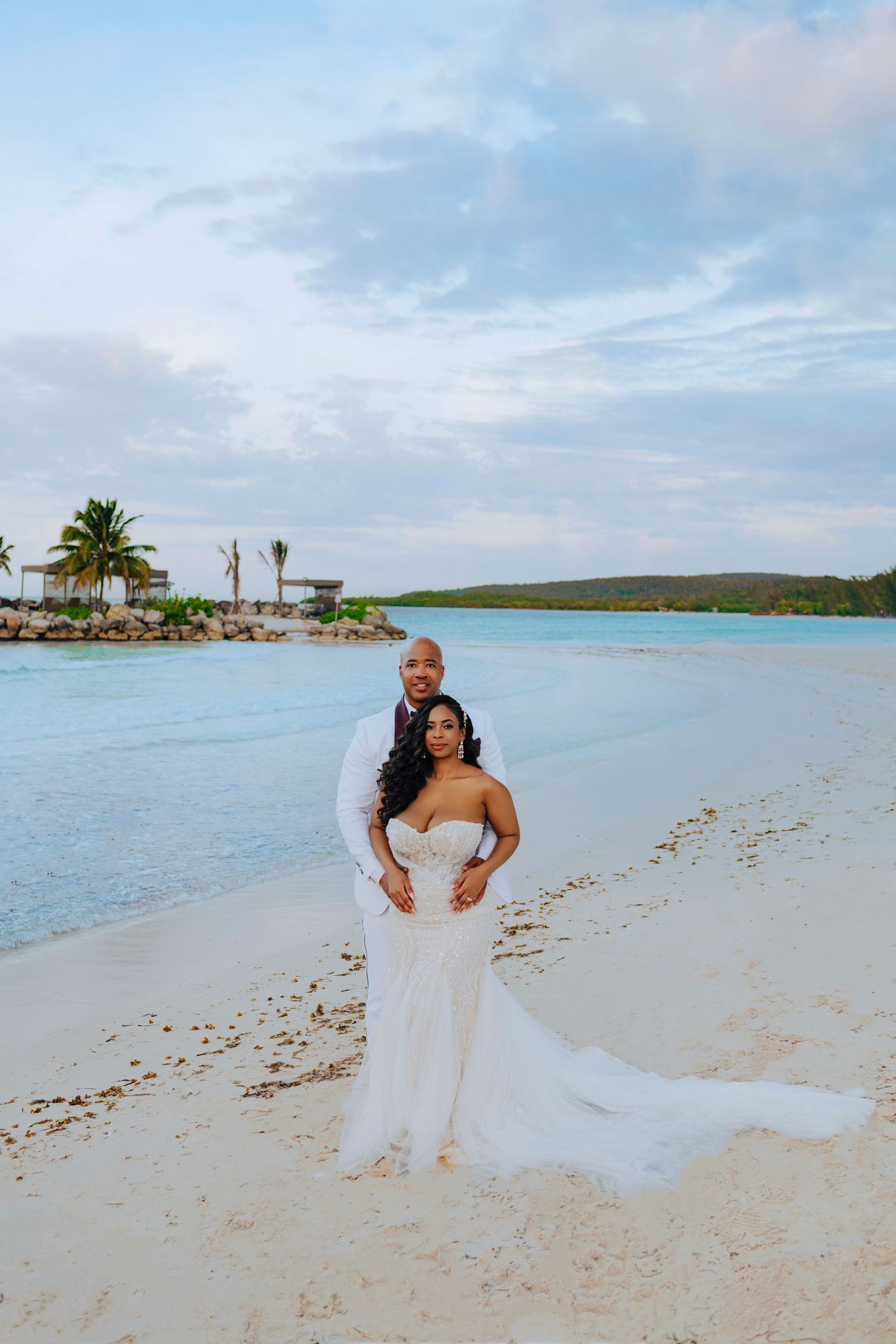 A couple stands together on a white sandy beach with turquoise waters and palm trees in the Caribbean background.